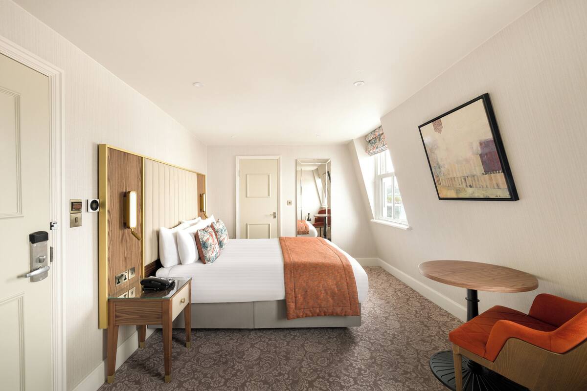 Une chambre d’hôtel avec un lit, un bureau, une chaise, une petite table et un tableau au mur.