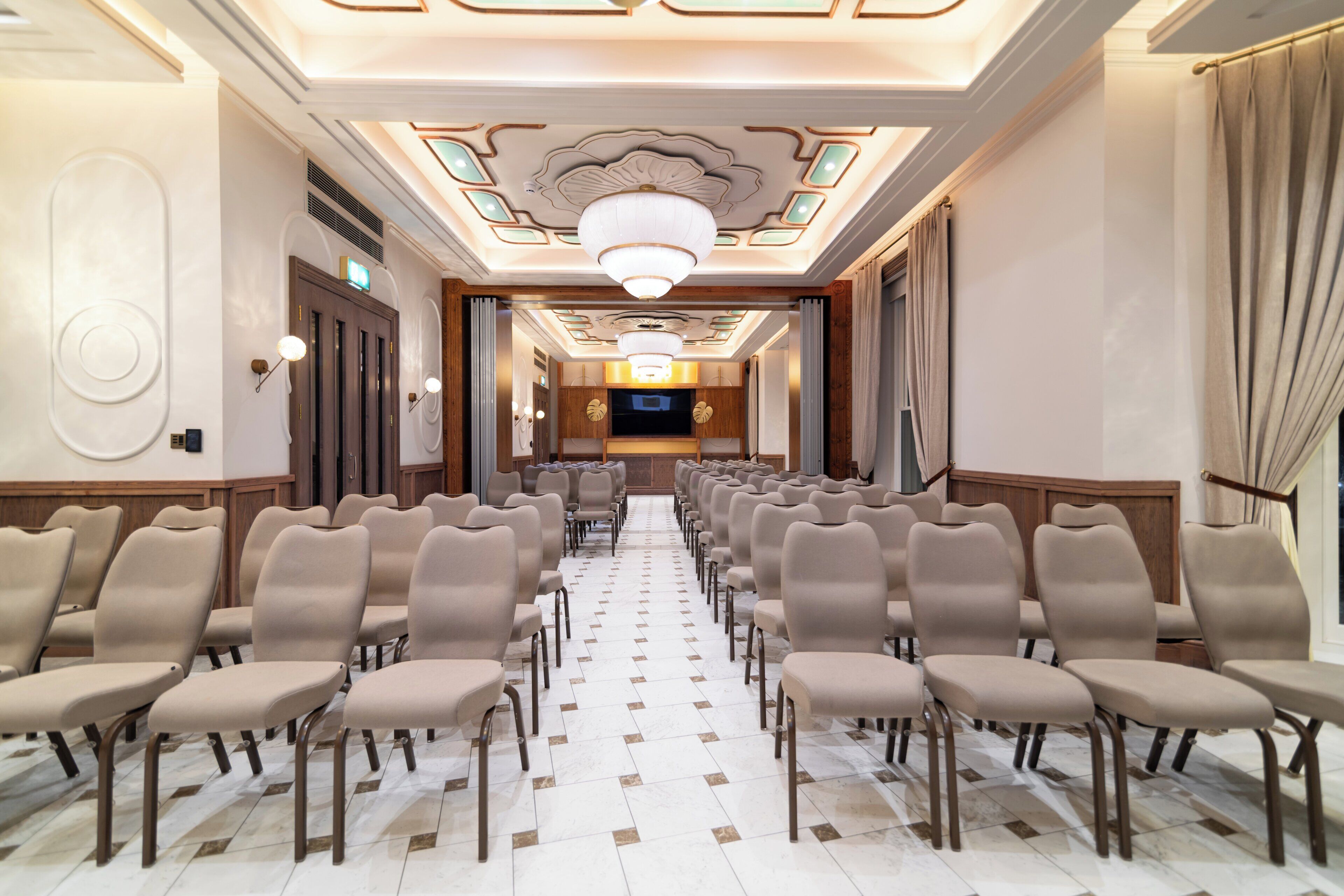A hotel lobby with rows of chairs, a chandelier, and a television screen.