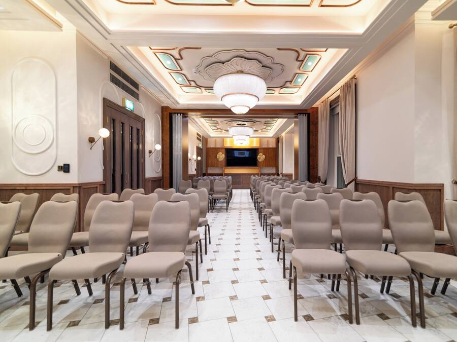 A hotel lobby with rows of chairs, a chandelier, and a television screen.