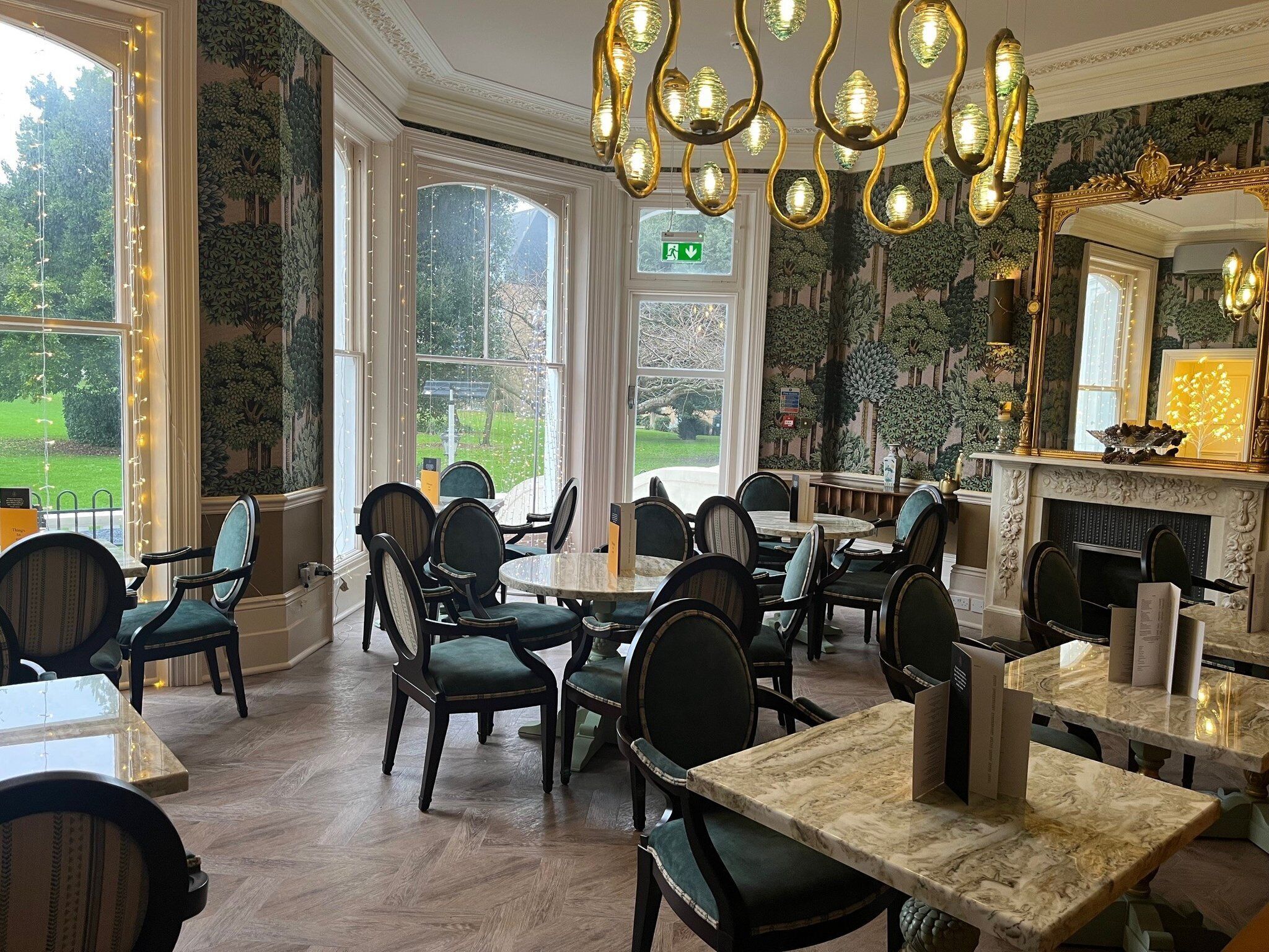 A dining area with marble tables and chairs, large windows with a view of greenery, and a fireplace with a mirror above it.