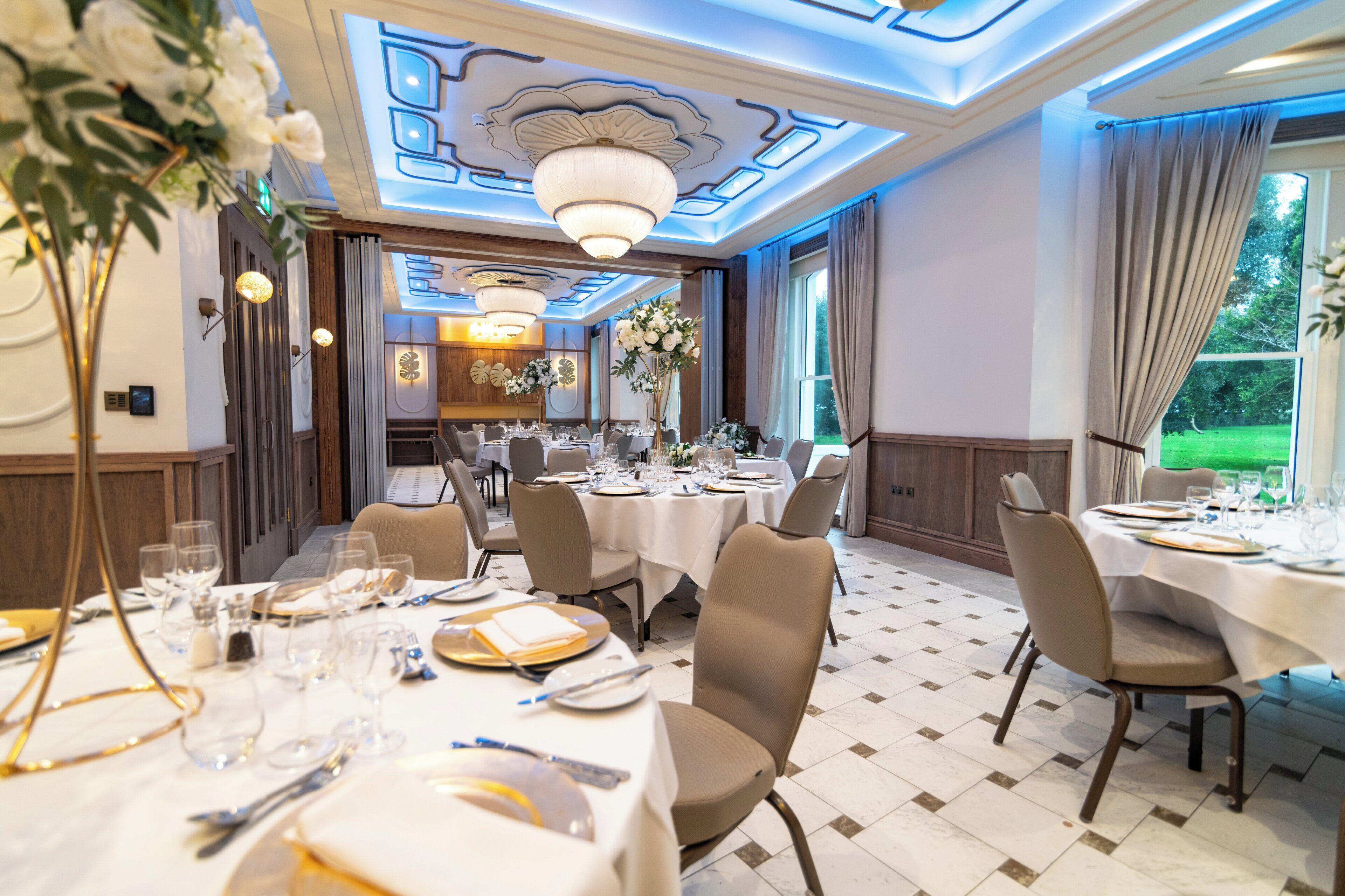 A formal dining room with multiple tables set for a meal, elegant chandeliers, and a view of the outdoors.