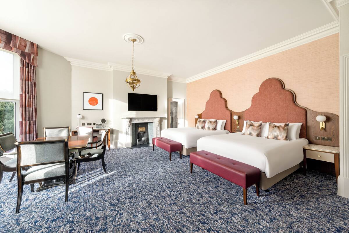 Une chambre d’hôtel avec deux lits, une cheminée, un coin repas avec des chaises et un téléviseur fixé au mur.