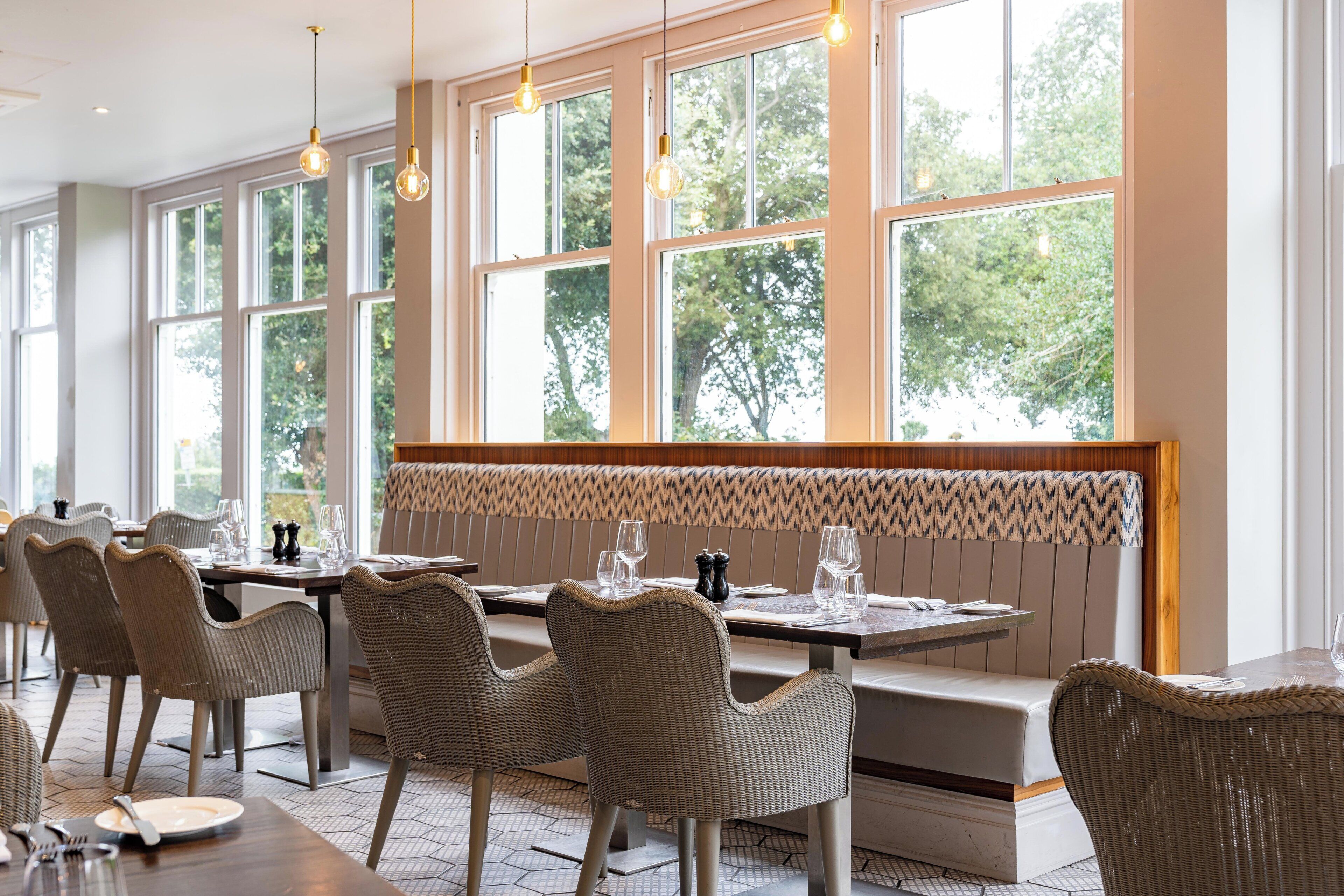 A restaurant with large windows, wicker chairs, and neatly set tables.