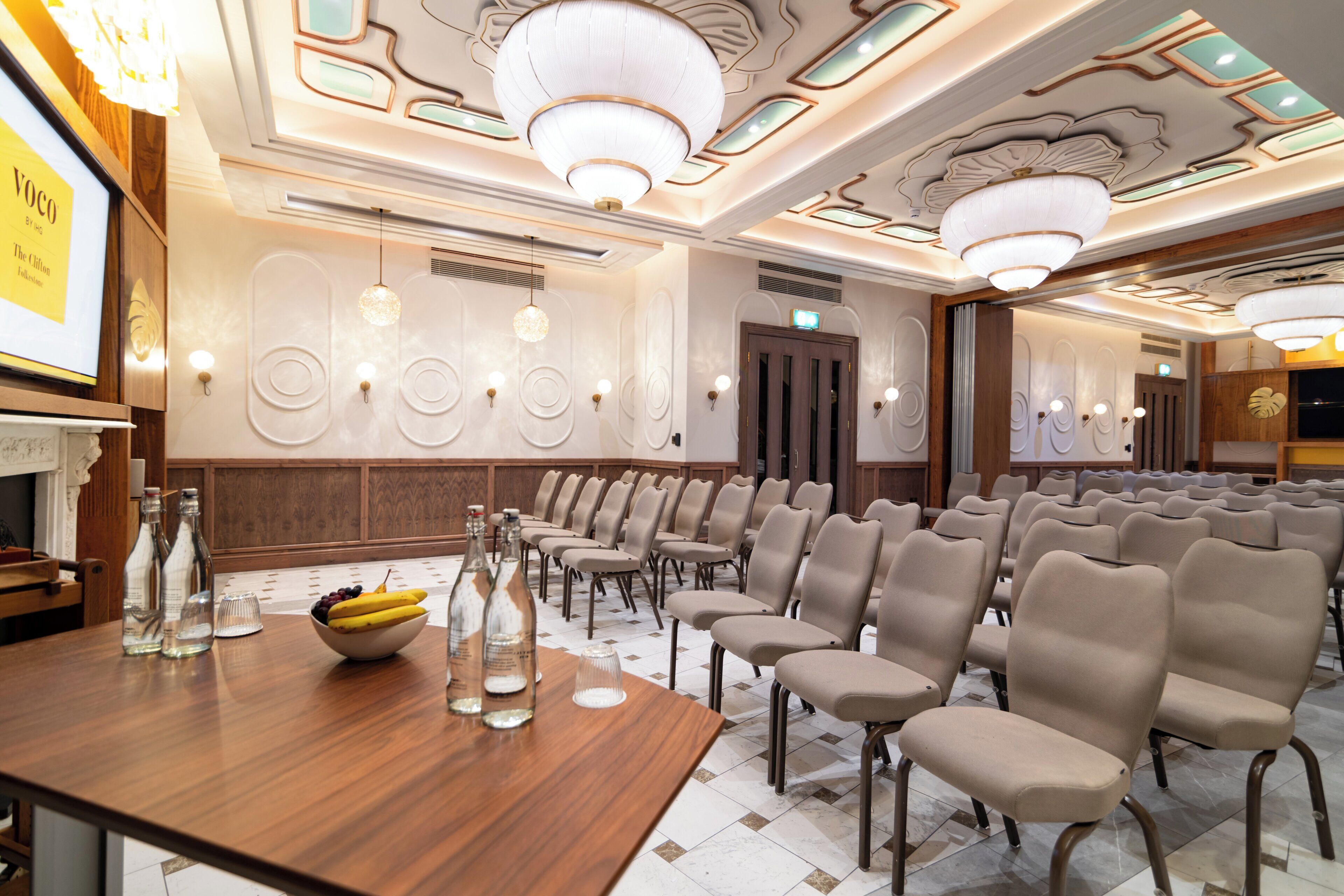 A conference room with rows of chairs, a wooden table with bottles and a bowl of fruit, and a chandelier.