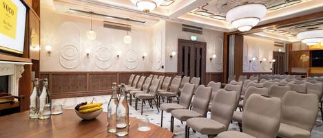 A conference room with rows of chairs, a wooden table with bottles and a bowl of fruit, and a chandelier.