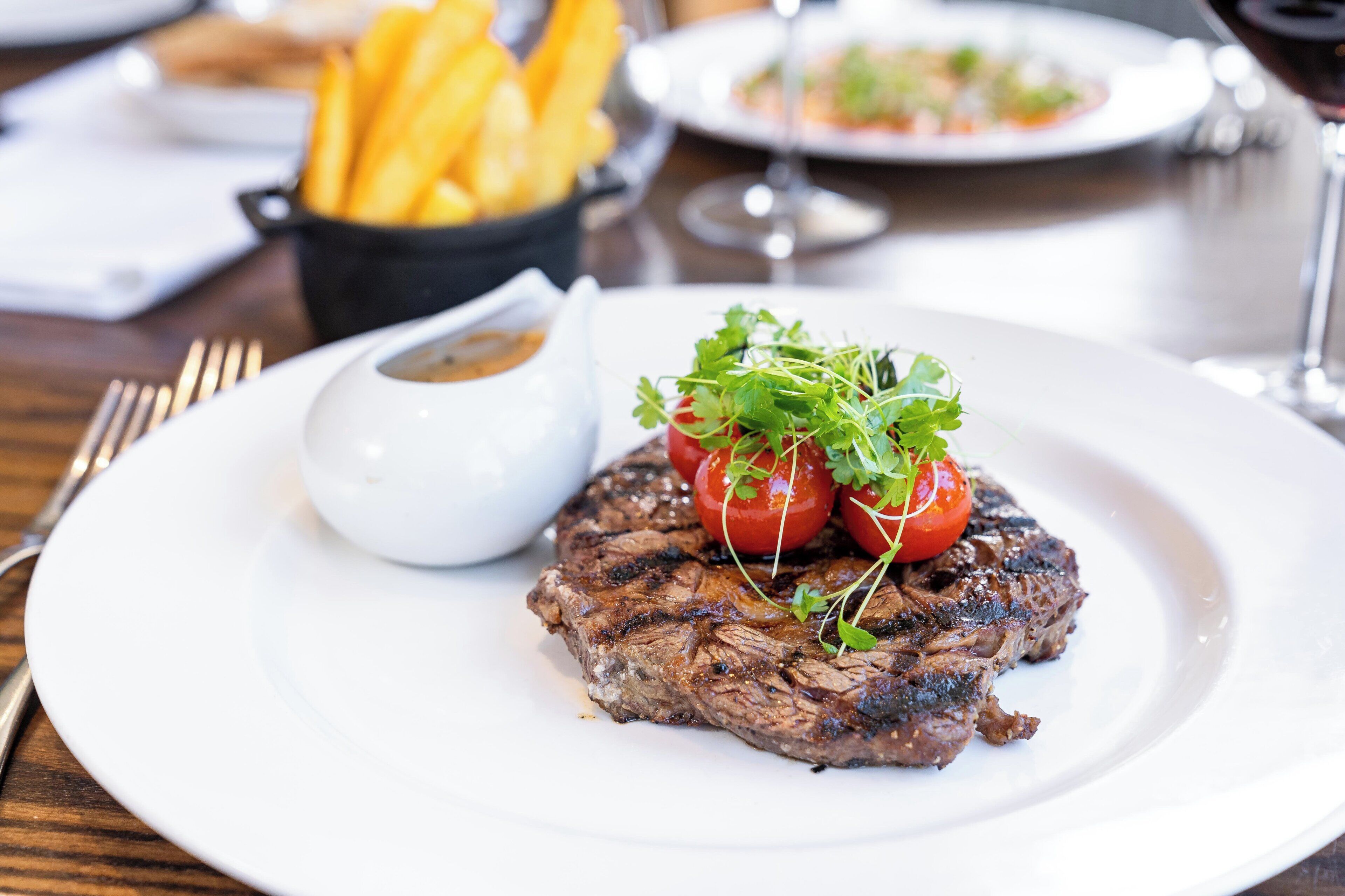Un bistec acompañado de papas fritas y un recipiente pequeño con salsa sobre un plato blanco.