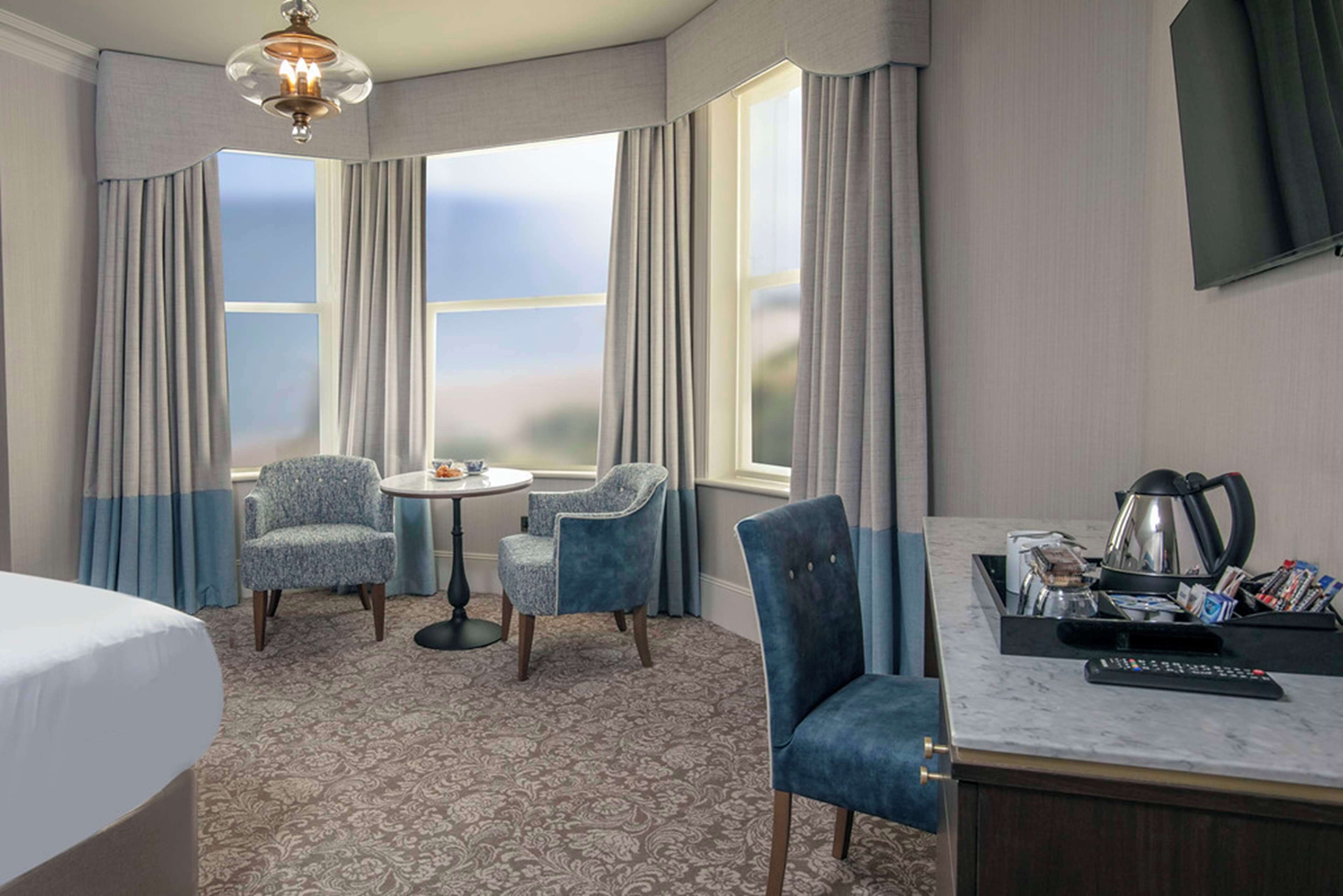 A hotel room with a bed, two armchairs, a small round table, a desk with a kettle, and a TV on the wall.