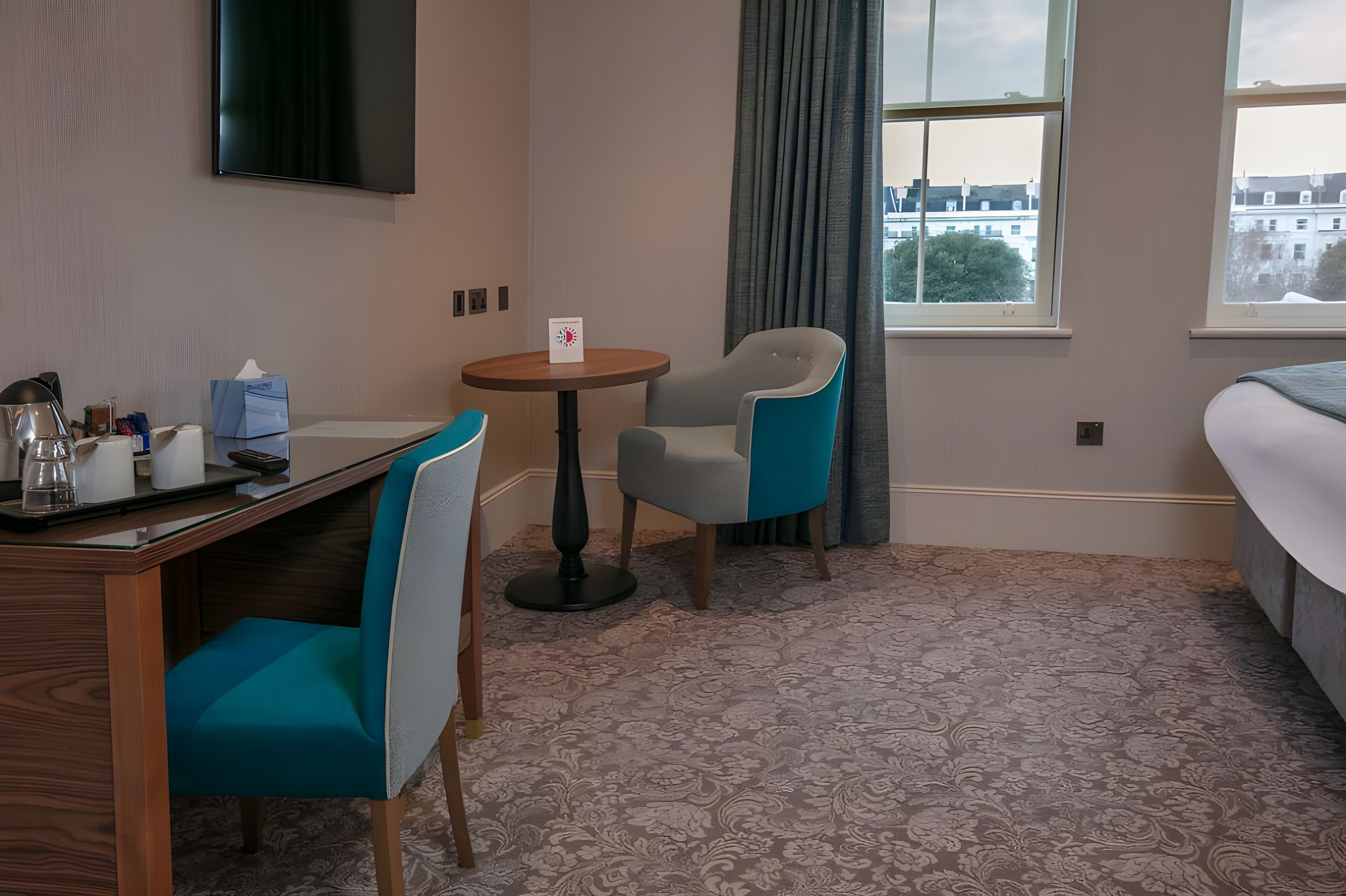 A hotel room with a bed, a desk with a chair, a small table with a chair, and a window with curtains.