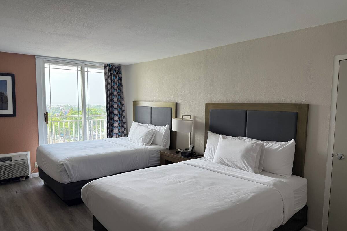 A hotel room with two beds, a window with a view, a bedside table with a lamp, and a radiator.