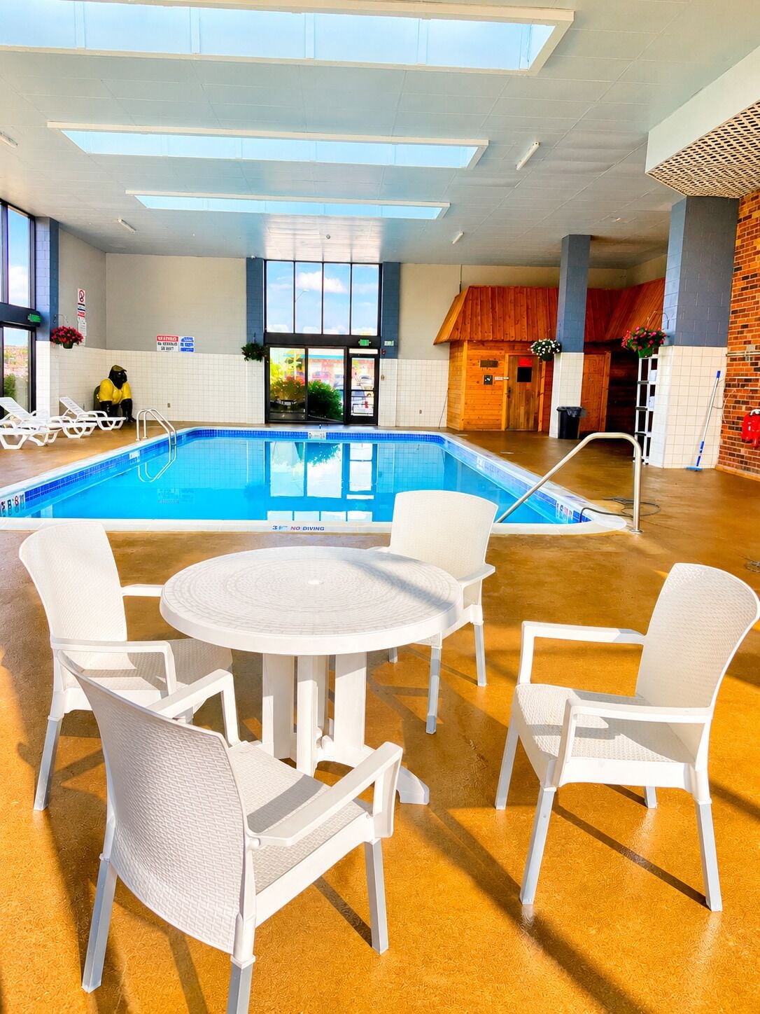 An indoor swimming pool with a table and chairs in the foreground, a wooden hut structure in the background, and a view of the outside through large windows.