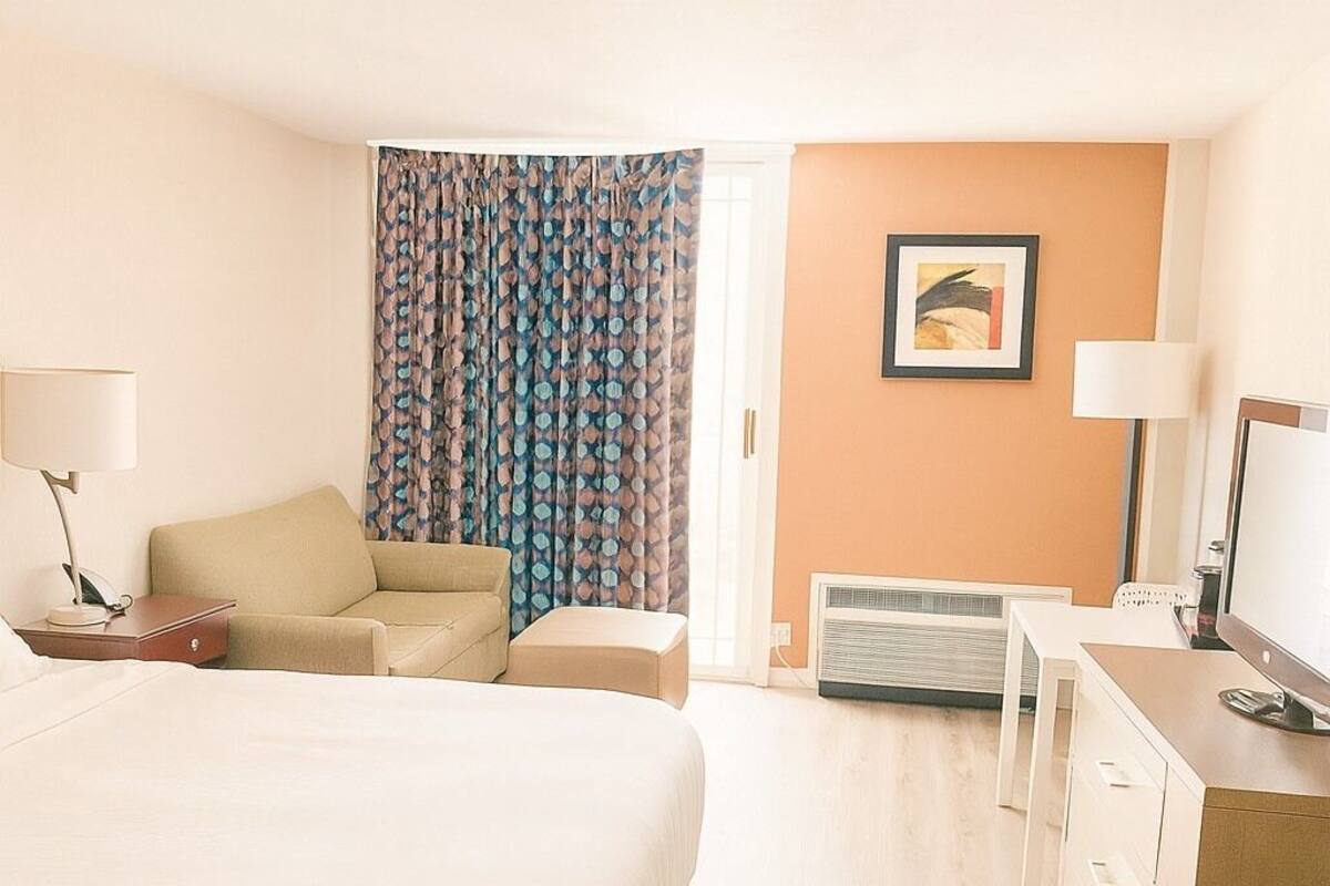 A hotel room with a large bed, a sofa, a desk with a computer, a TV, and a window with curtains.