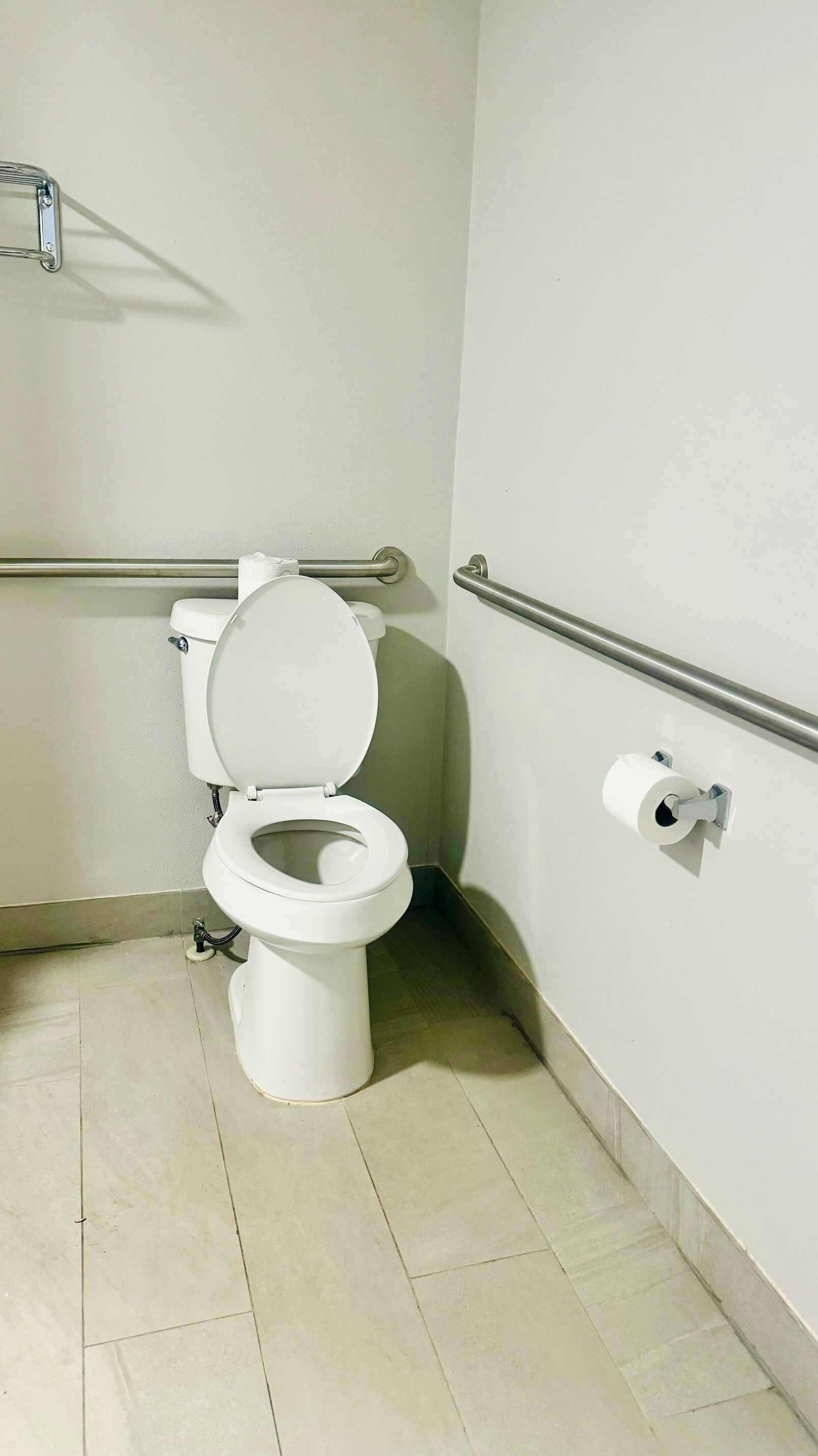 A standard white toilet with a metal grab bar, a roll of toilet paper on the wall, and a tiled floor.