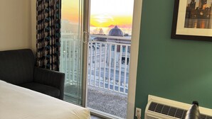 Habitación de hotel con cama, una silla, vista a un paisaje urbano al atardecer, un cuadro en la pared y una ventana con cortina.