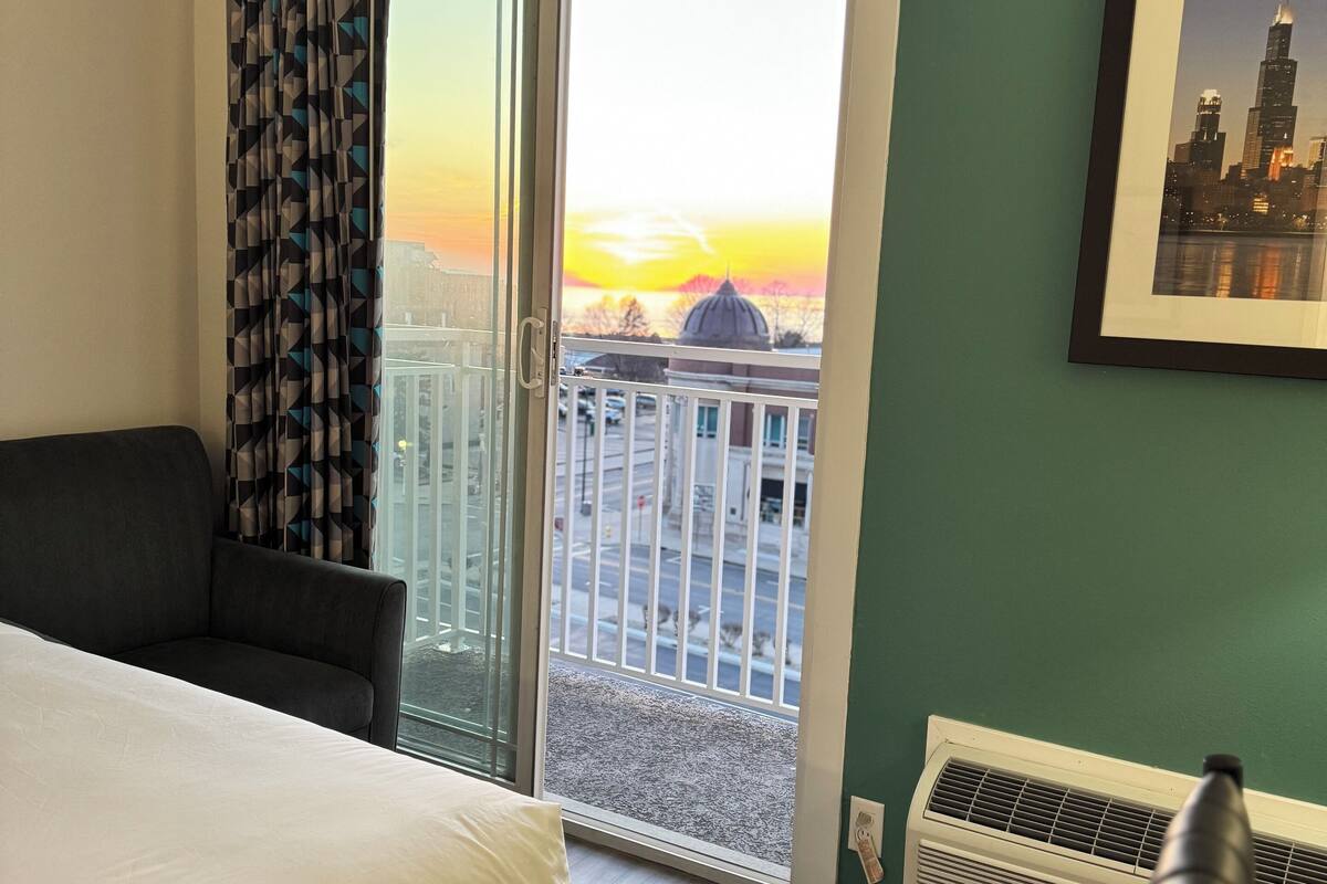 A hotel room with a bed, a chair, a view of a cityscape at sunset, a framed picture on the wall, and a window with a curtain.