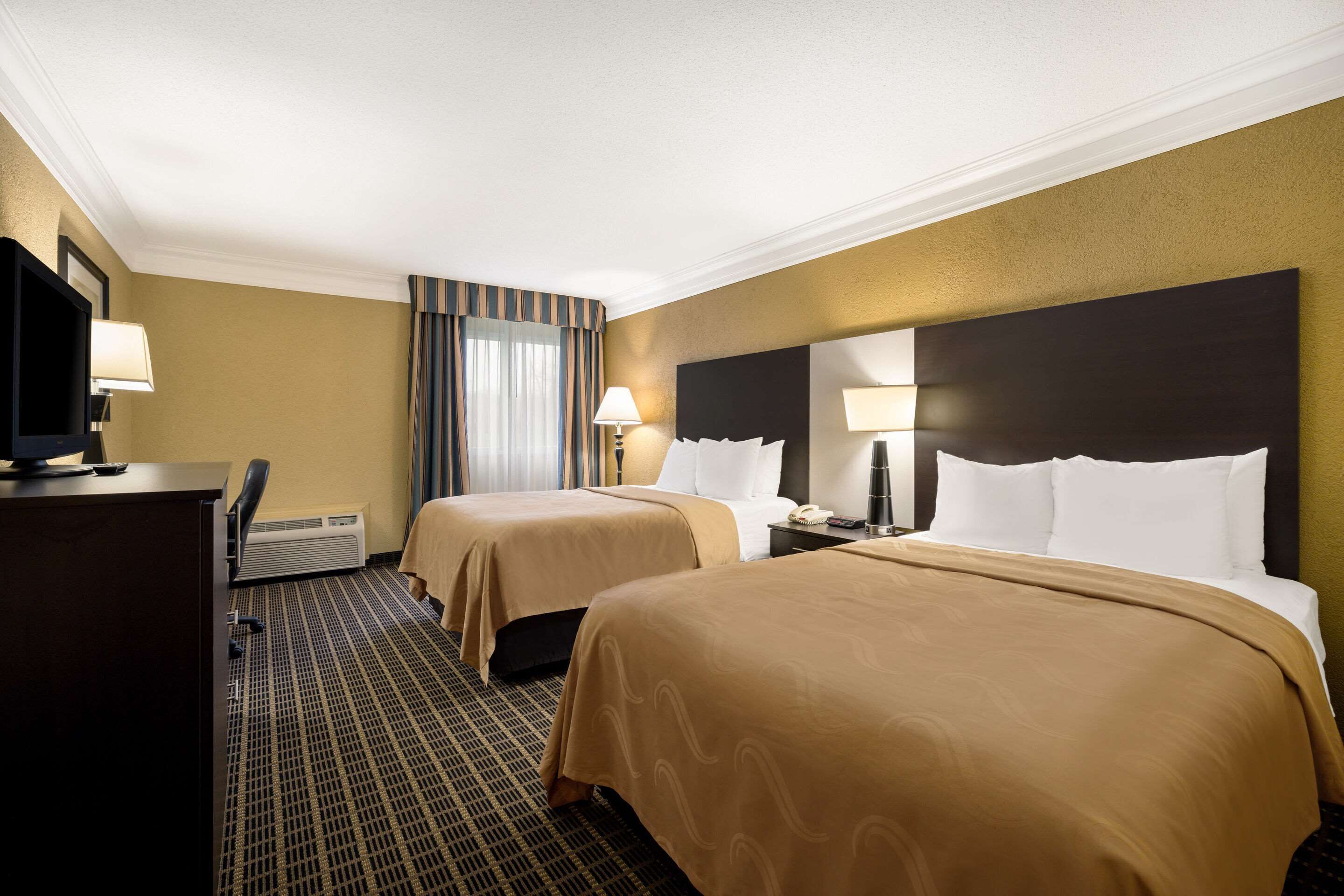 A hotel room with two beds, a desk, a television, and a window with curtains.