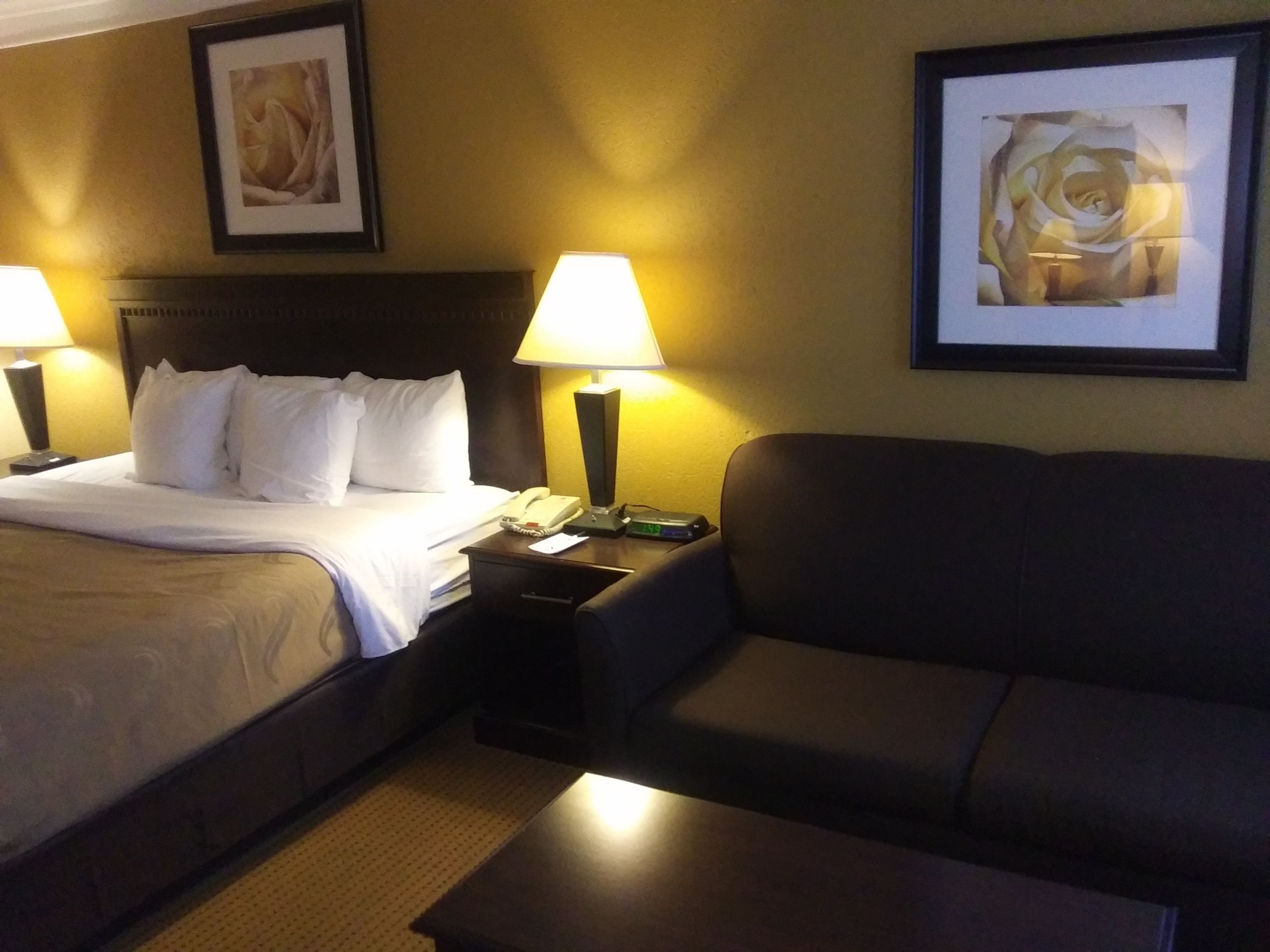 A hotel room with a bed, a sofa, two bedside lamps, a nightstand with a phone and a clock, and two framed artworks on the wall.
