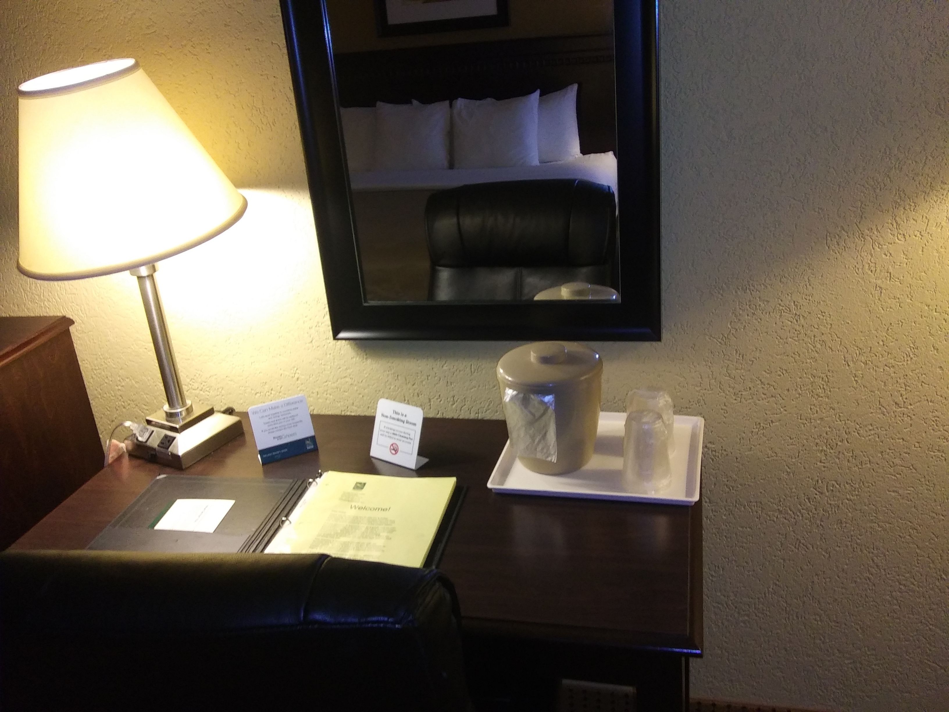A hotel room with a bedside lamp, a mirror, a bedside table with a laptop, a welcome card, a coffee maker, and a tray with sugar and cream containers.