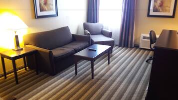 A hotel room with a sofa, armchair, and a small table. There is a lamp on a side table, and a framed picture on the wall.