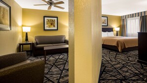 Une chambre d’hôtel avec un ventilateur de plafond, un canapé, un lit recouvert d’une courtepointe beige, une moquette à motifs et des œuvres d’art encadrées accrochées aux murs.