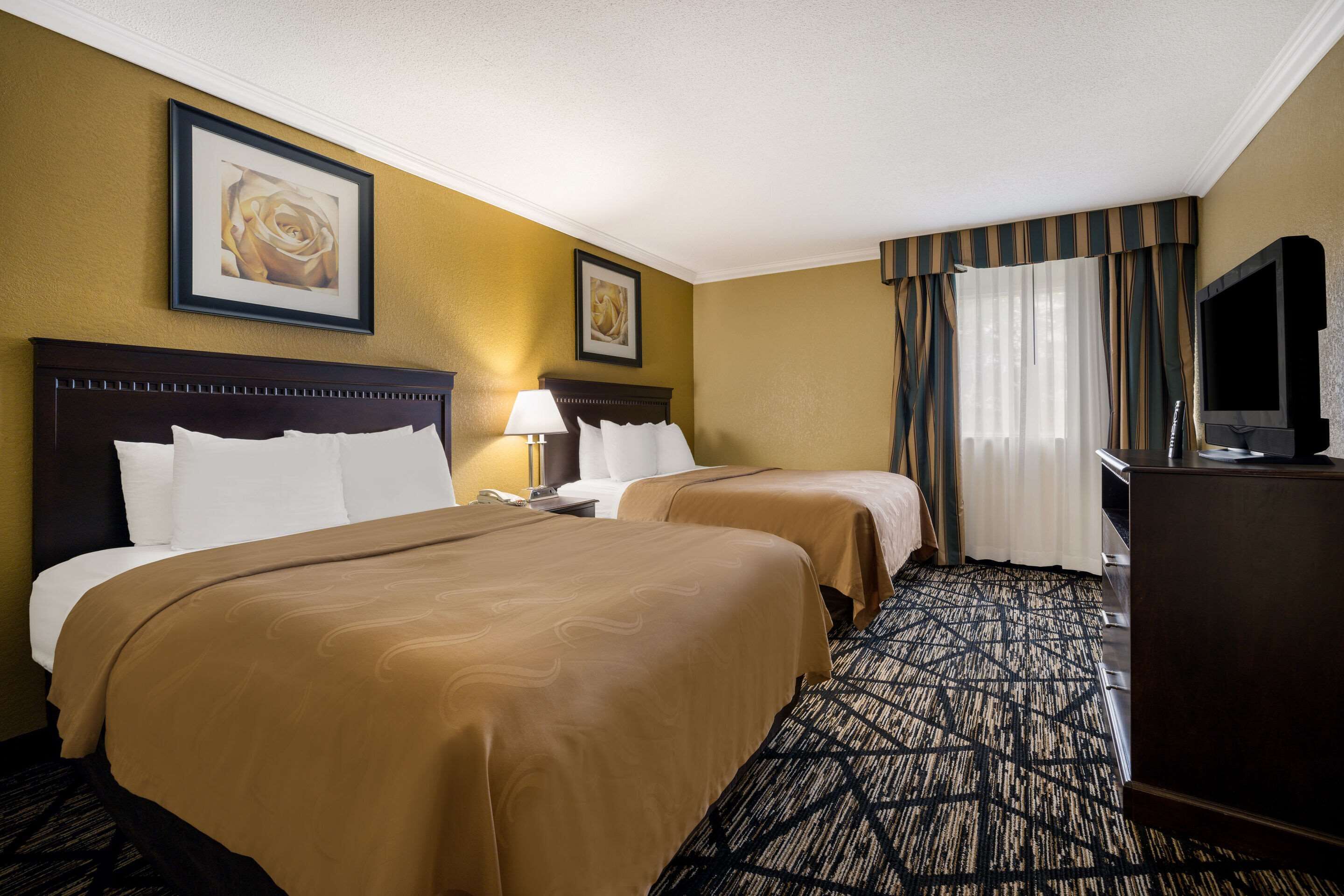 Quarto de hotel com duas camas, uma televisão, uma escrivaninha e um abajur.