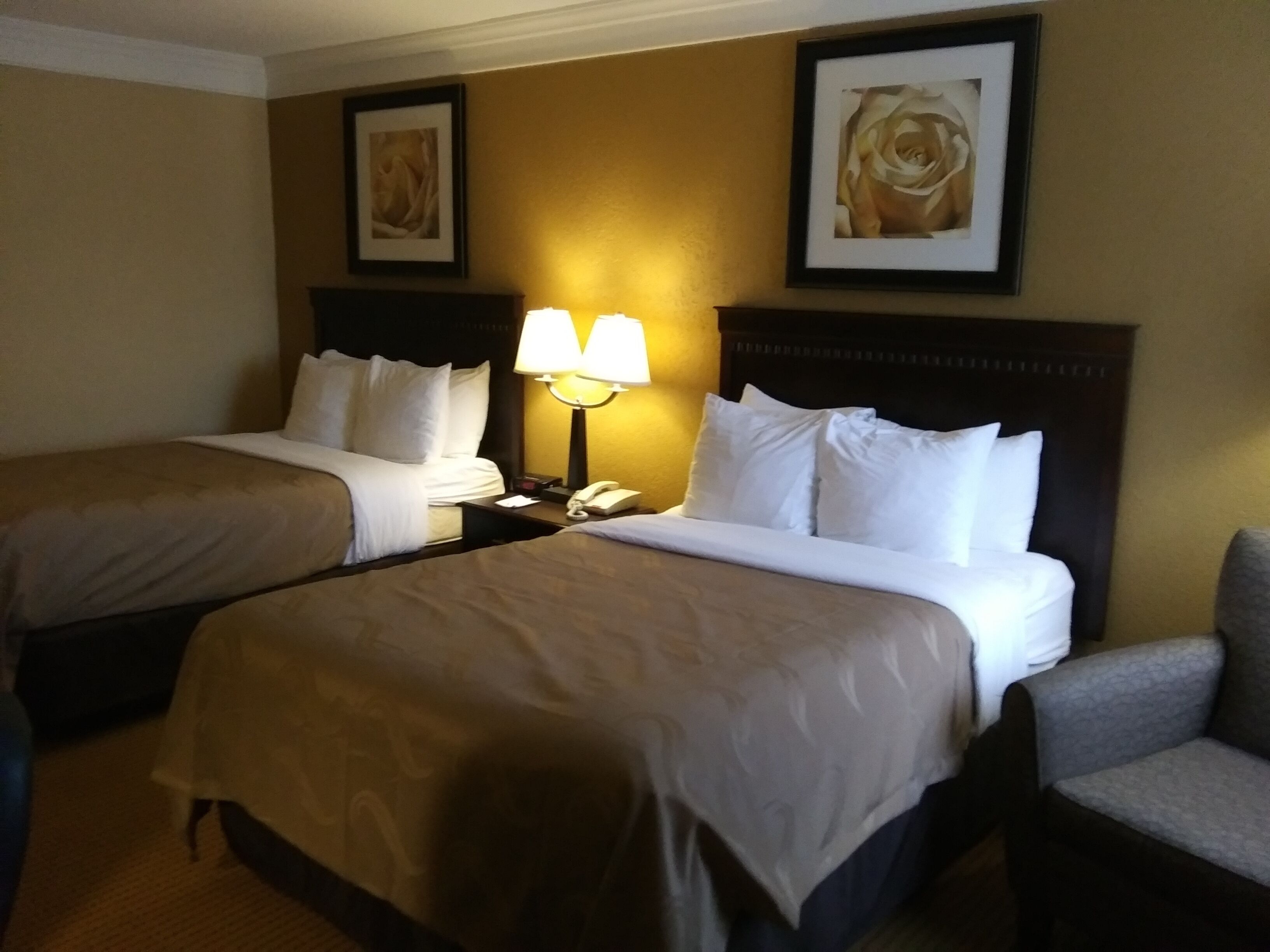 Ein Hotelzimmer mit zwei Betten, einem Nachttisch mit Lampe und gerahmten Kunstwerken an den Wänden.