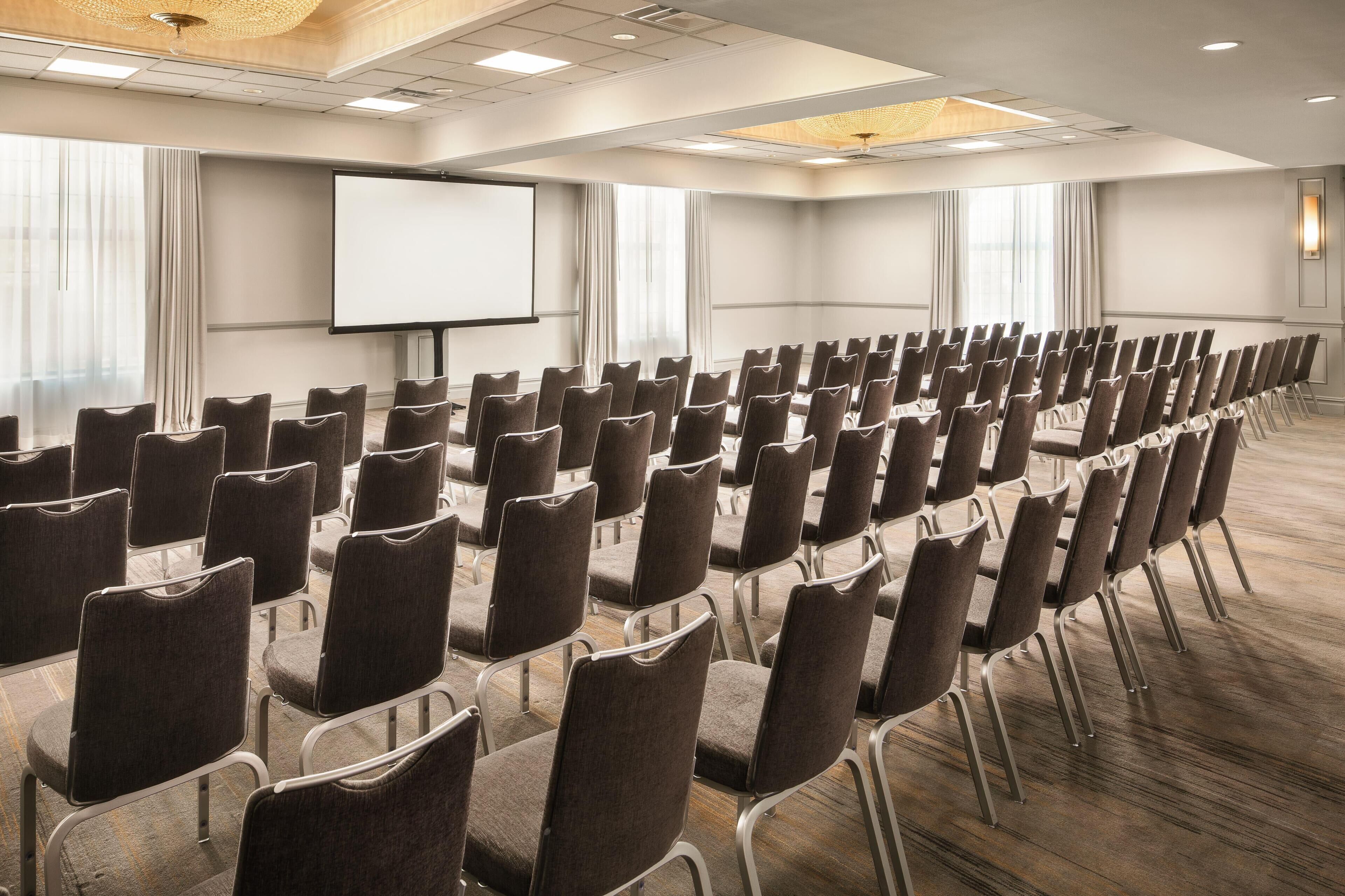 Un salón de conferencias con filas de sillas, una pantalla de proyección y amplias ventanas.