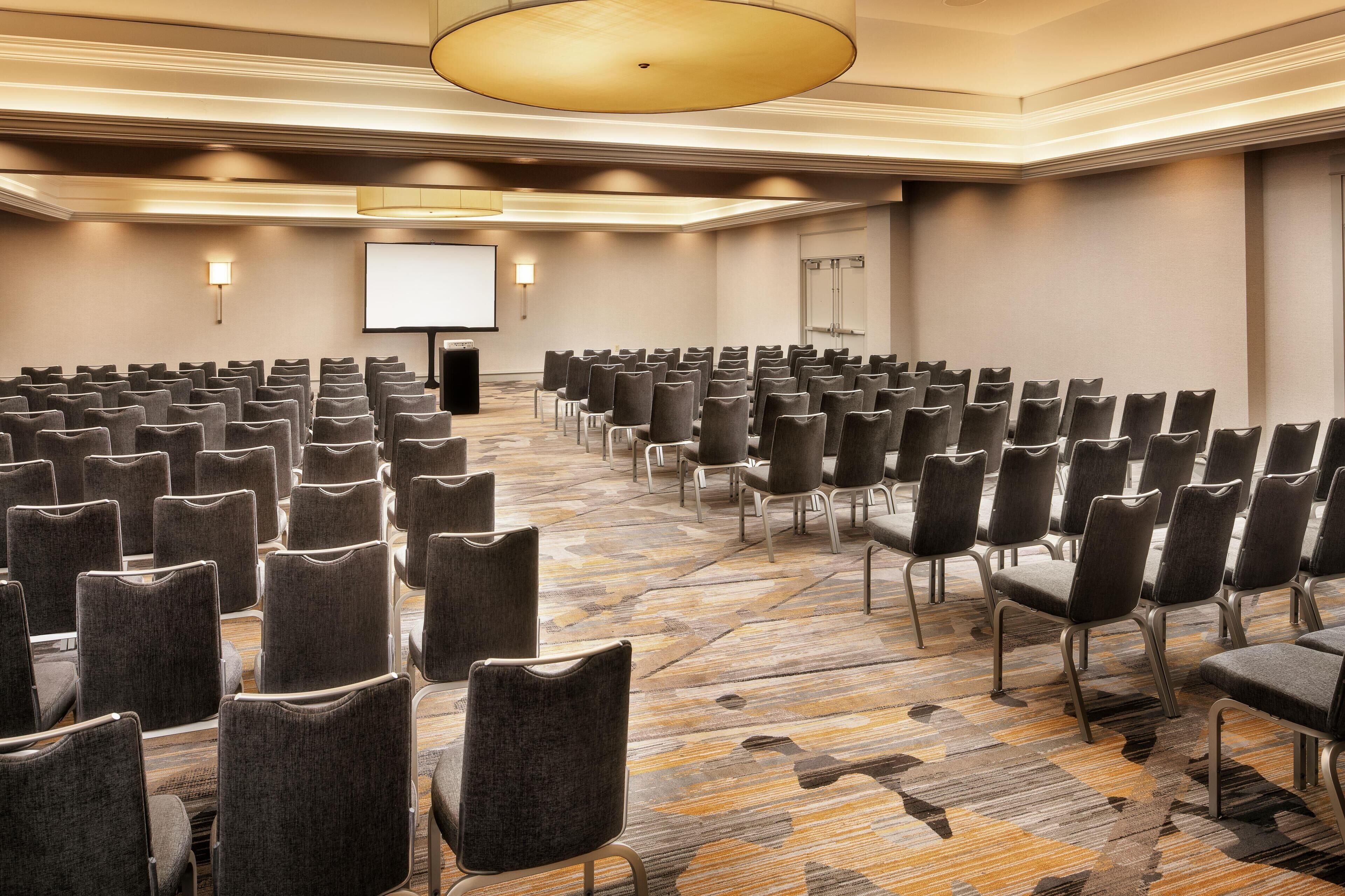 A conference room with rows of chairs, a projector screen, and a carpeted floor.