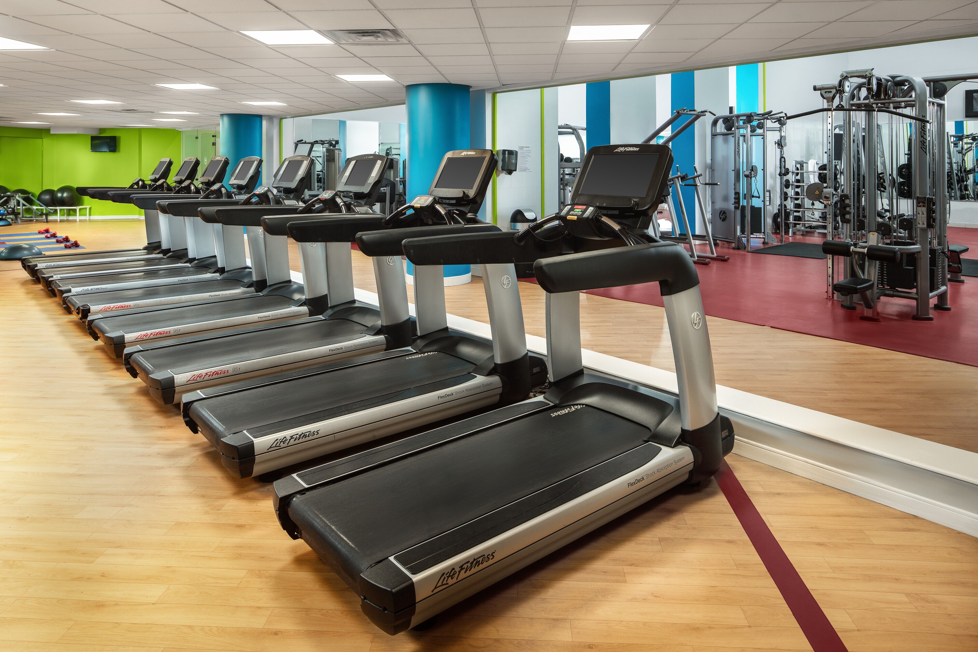Eine Reihe von Laufbändern in einem Fitnessstudio mit Spiegelwand und verschiedenen Trainingsgeräten.