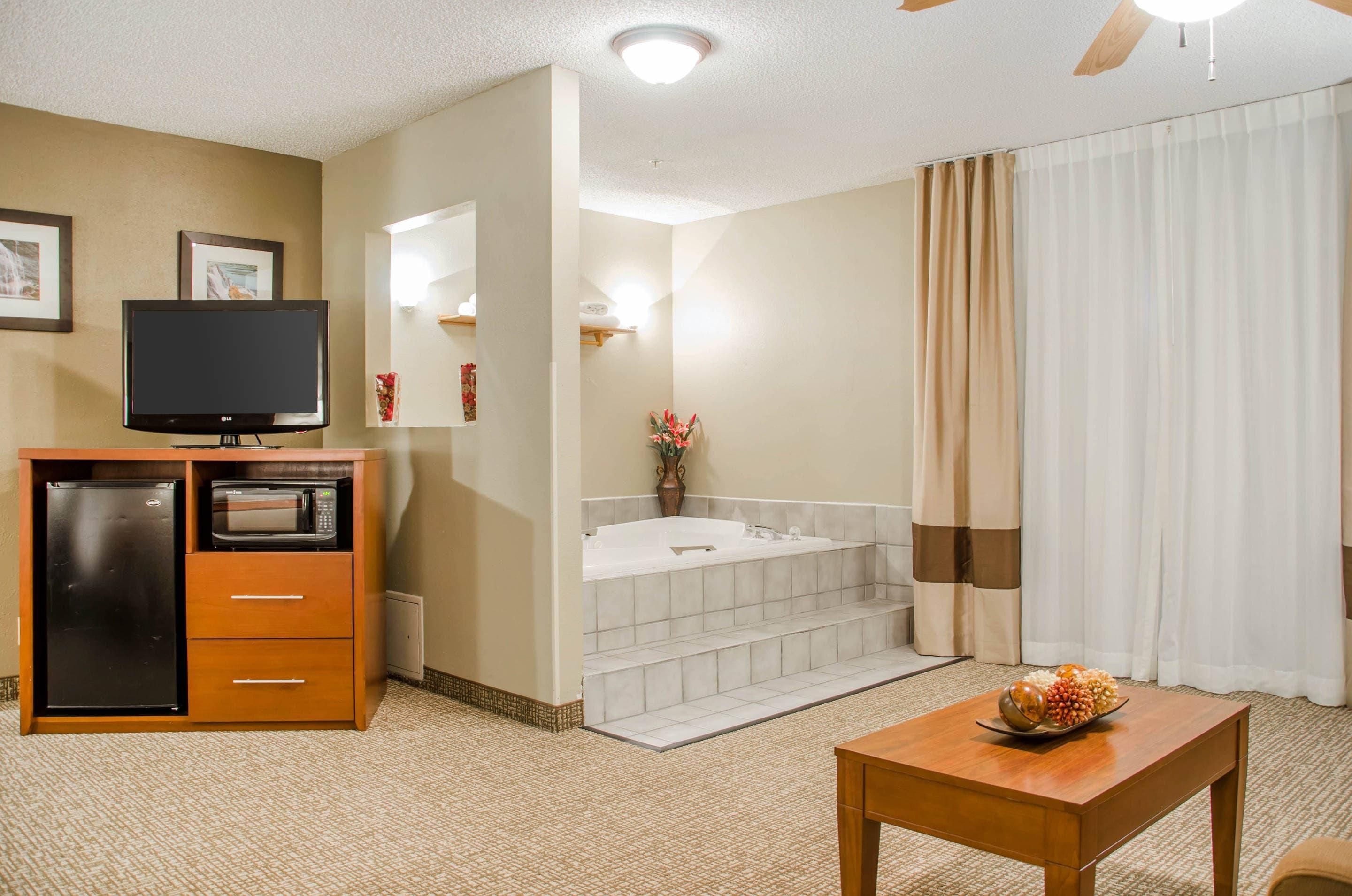 A hotel room with a television, a refrigerator, a microwave, a wooden table, and a bathroom with a bathtub and a shower curtain.