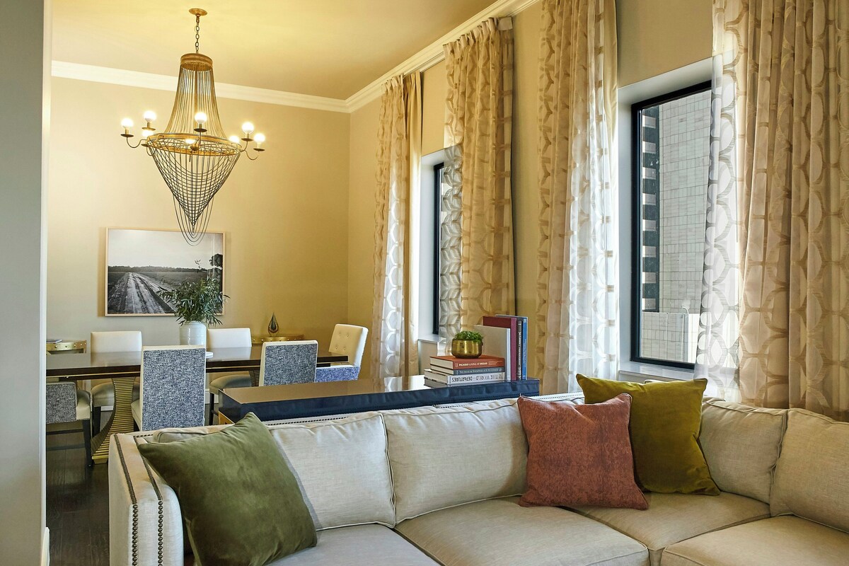 A living room with a sofa, a dining area with chairs, a chandelier, and a window with curtains.