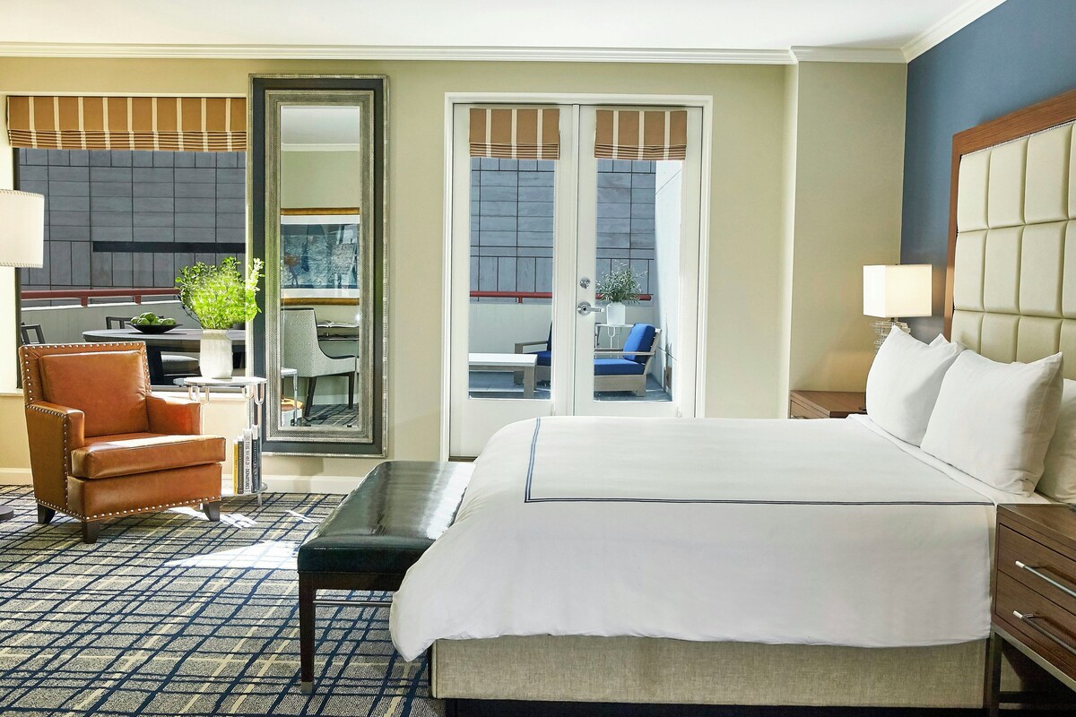A hotel room with a large bed, a desk, a chair, a mirror, and a view of the outside.