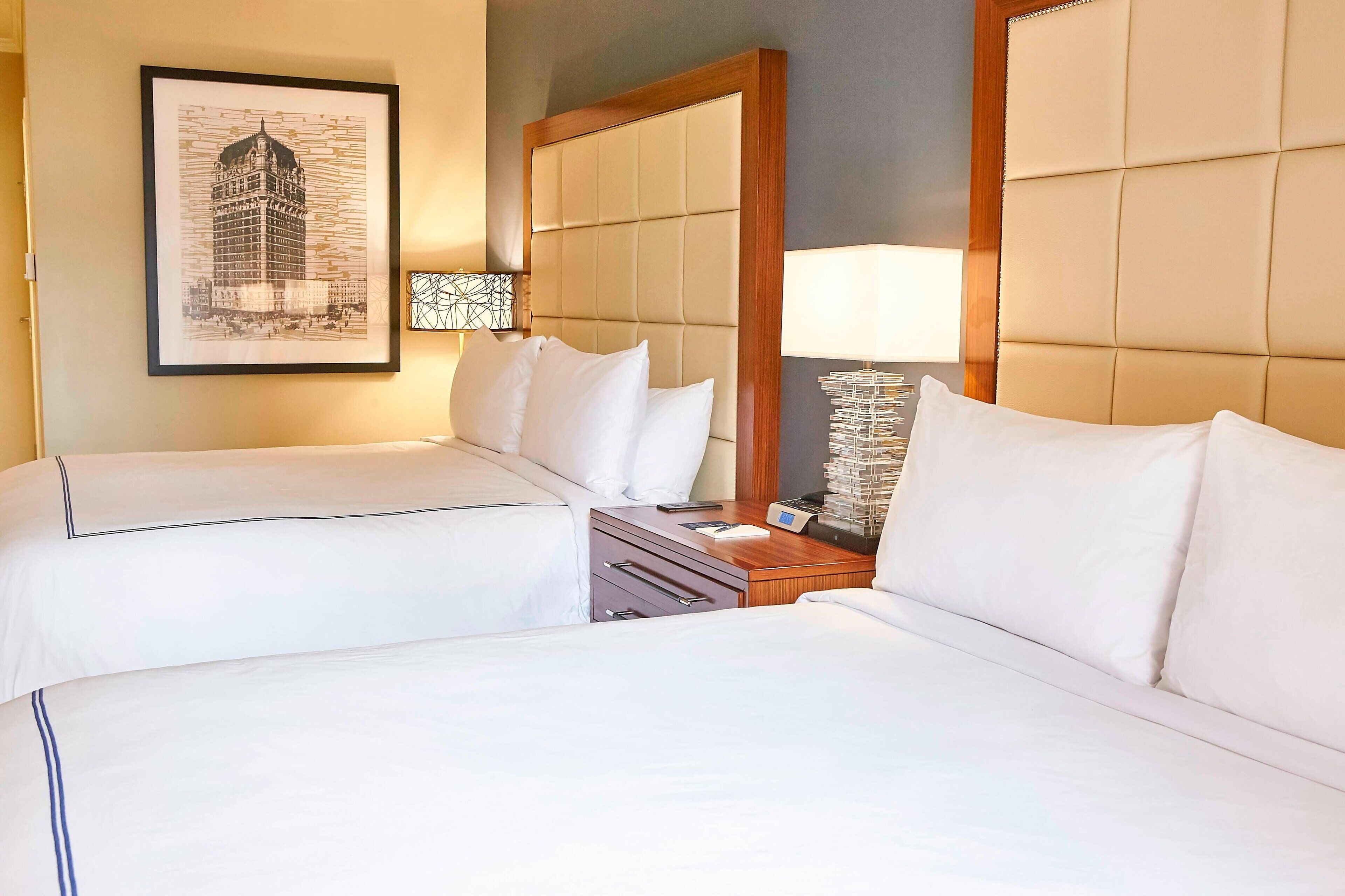 A hotel room with two beds, a large mirror, a bedside table with a lamp, and a framed picture on the wall.
