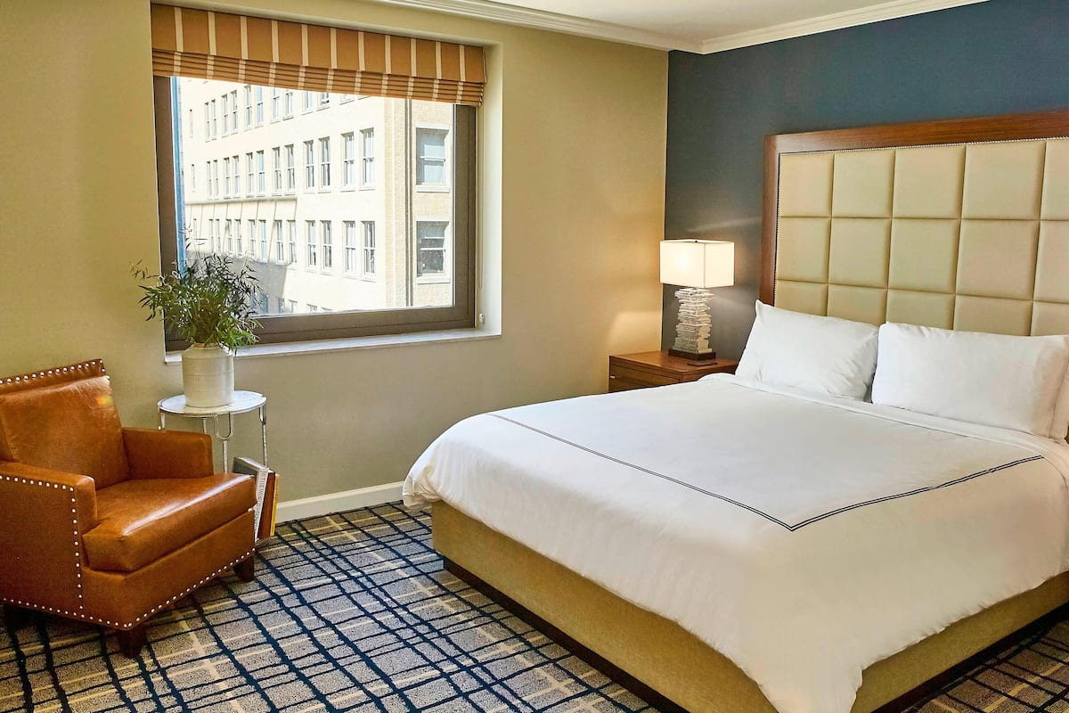 Habitación de hotel con una cama grande, una silla, una mesita con una lámpara y una ventana con vistas a edificios.