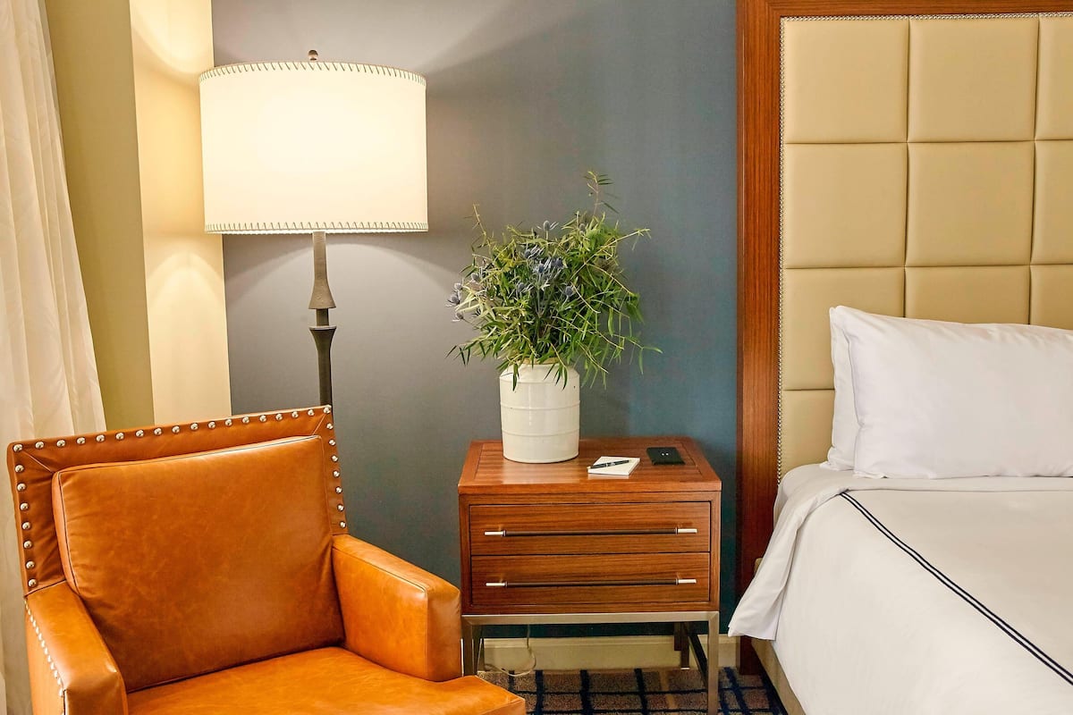 Habitación de hotel con una silla de cuero marrón, una mesita de noche con lámpara y planta en maceta, y una cama con ropa blanca.