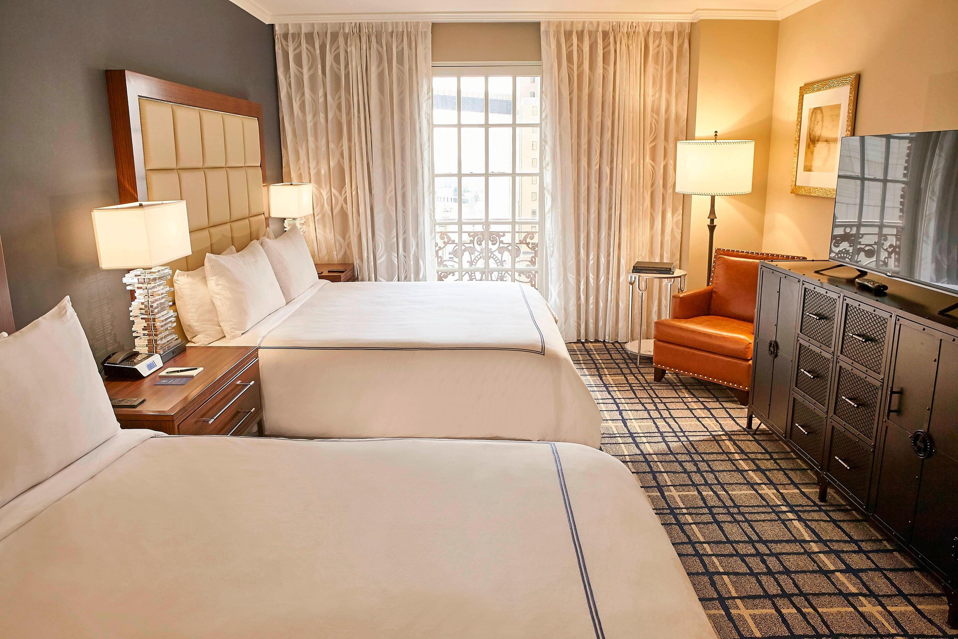 A hotel room with two beds, a desk with a lamp, a TV, a sofa, and a patterned carpet.