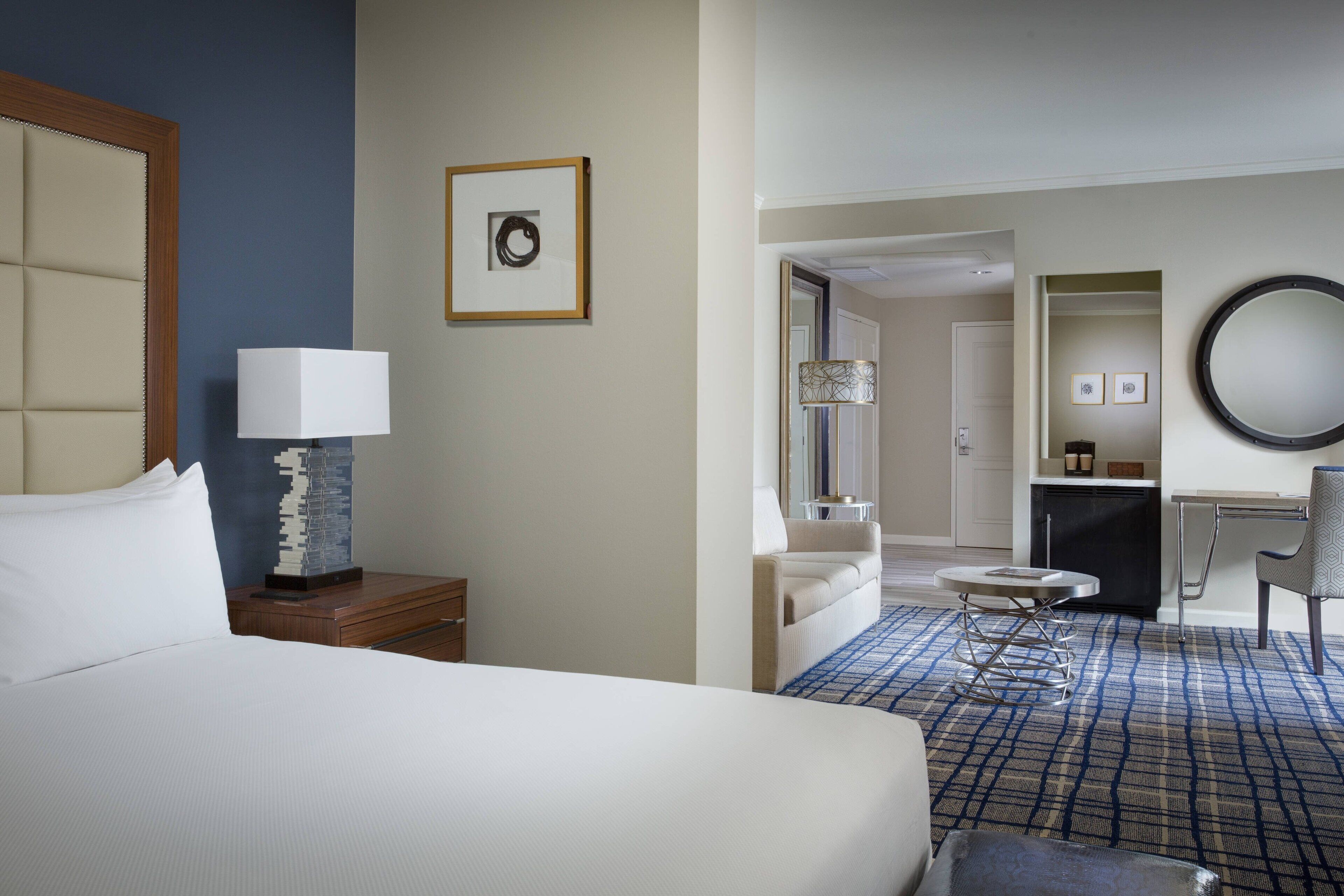 A hotel room with a bed, a desk, a chair, a lamp, a round mirror, and a small table.
