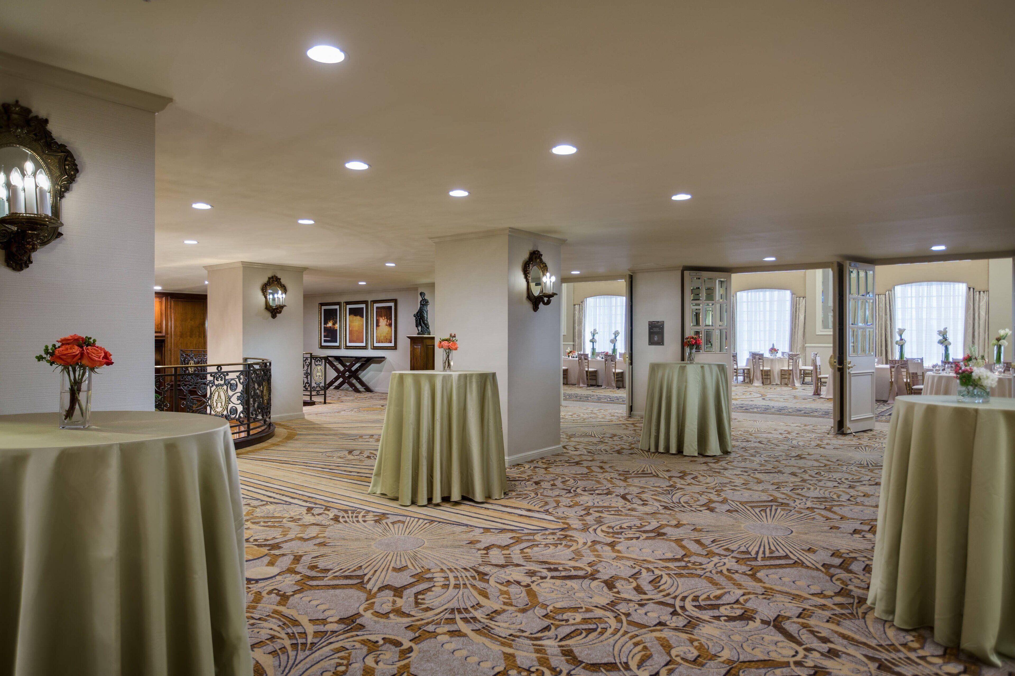 A spacious hotel lobby with multiple tables, ornate carpet, and a grand staircase.