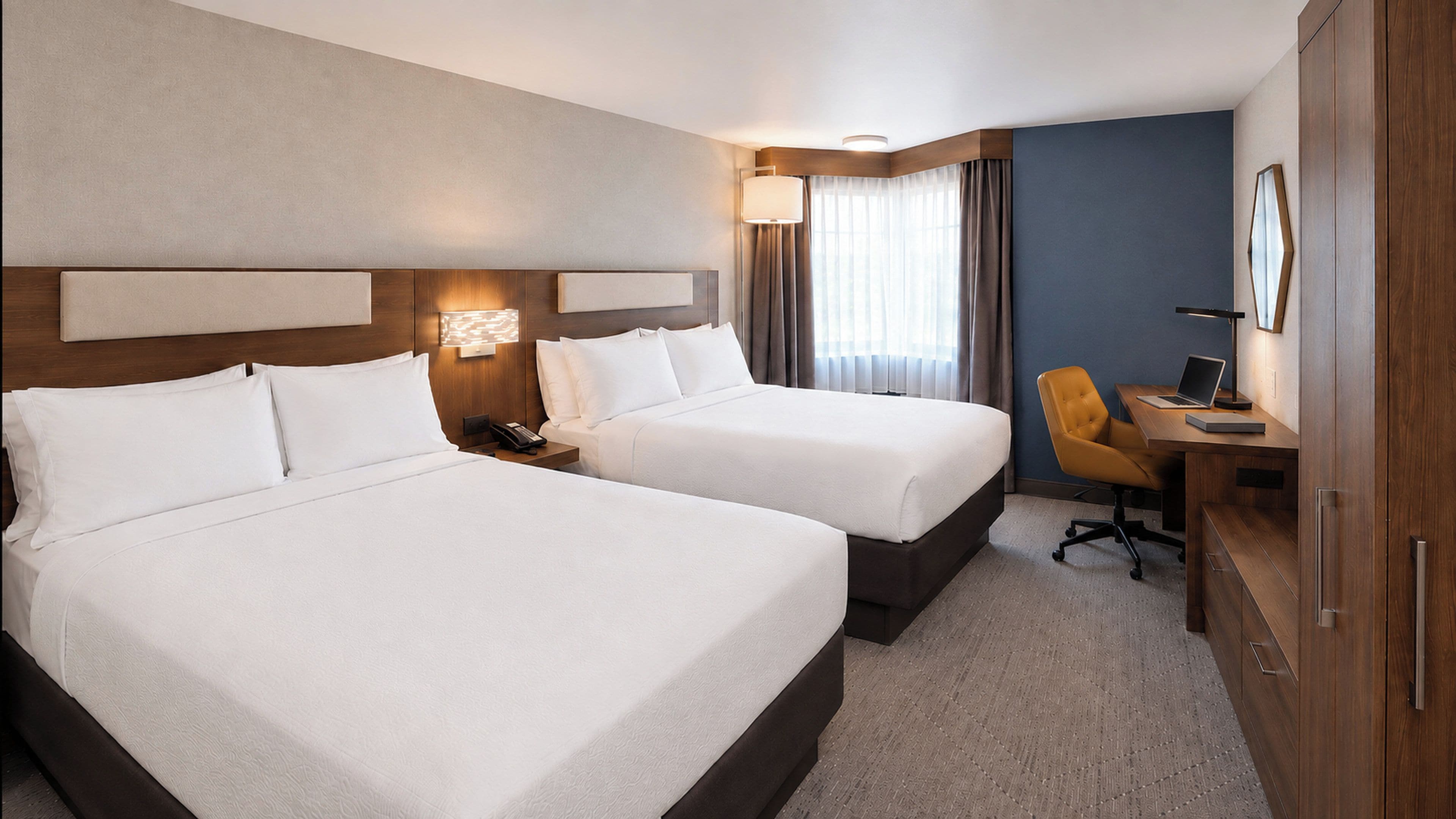 Habitación de hotel con dos camas, un escritorio con silla, una lámpara y un armario.