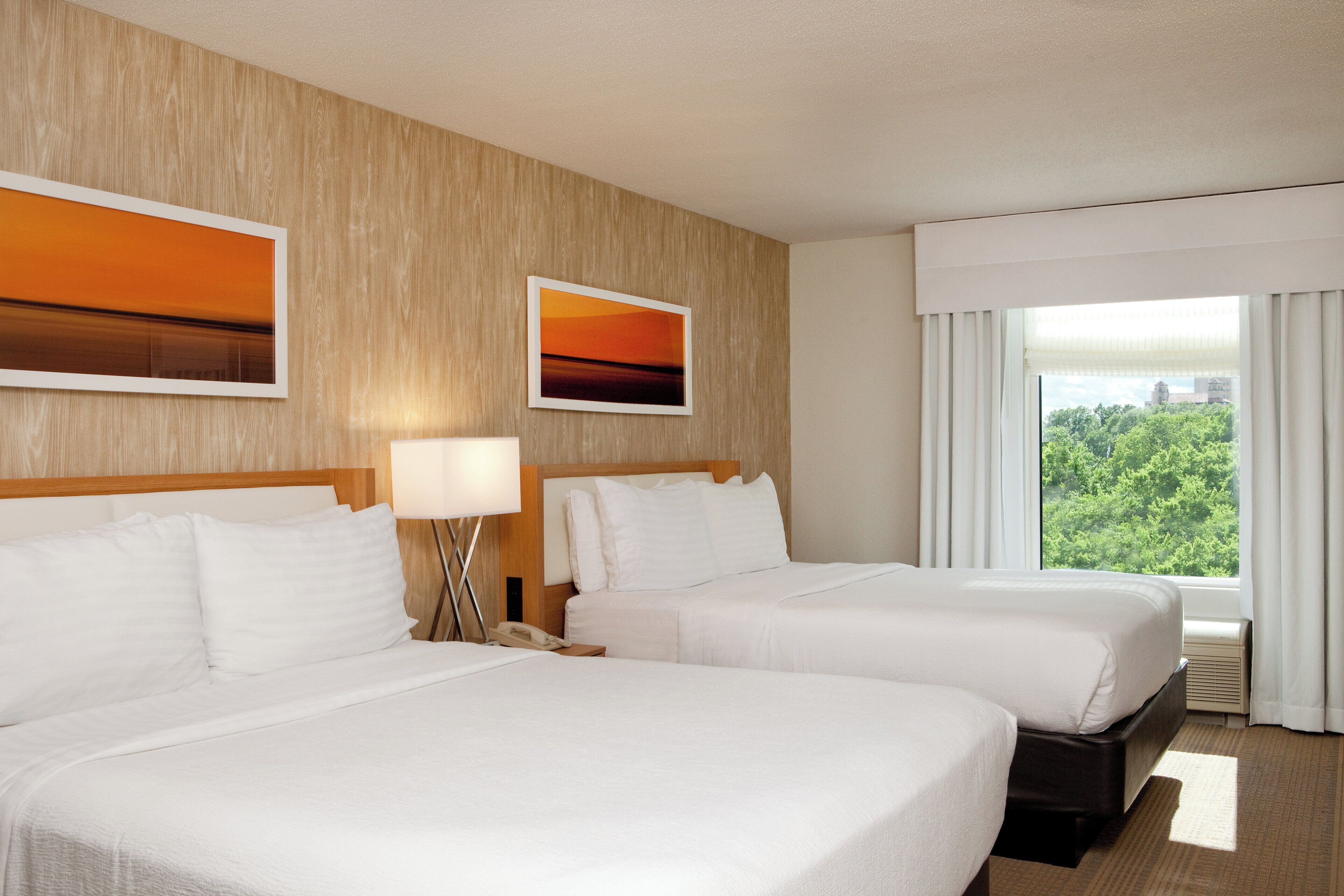 A hotel room with two beds, a nightstand, a lamp, and a window with a view of greenery.
