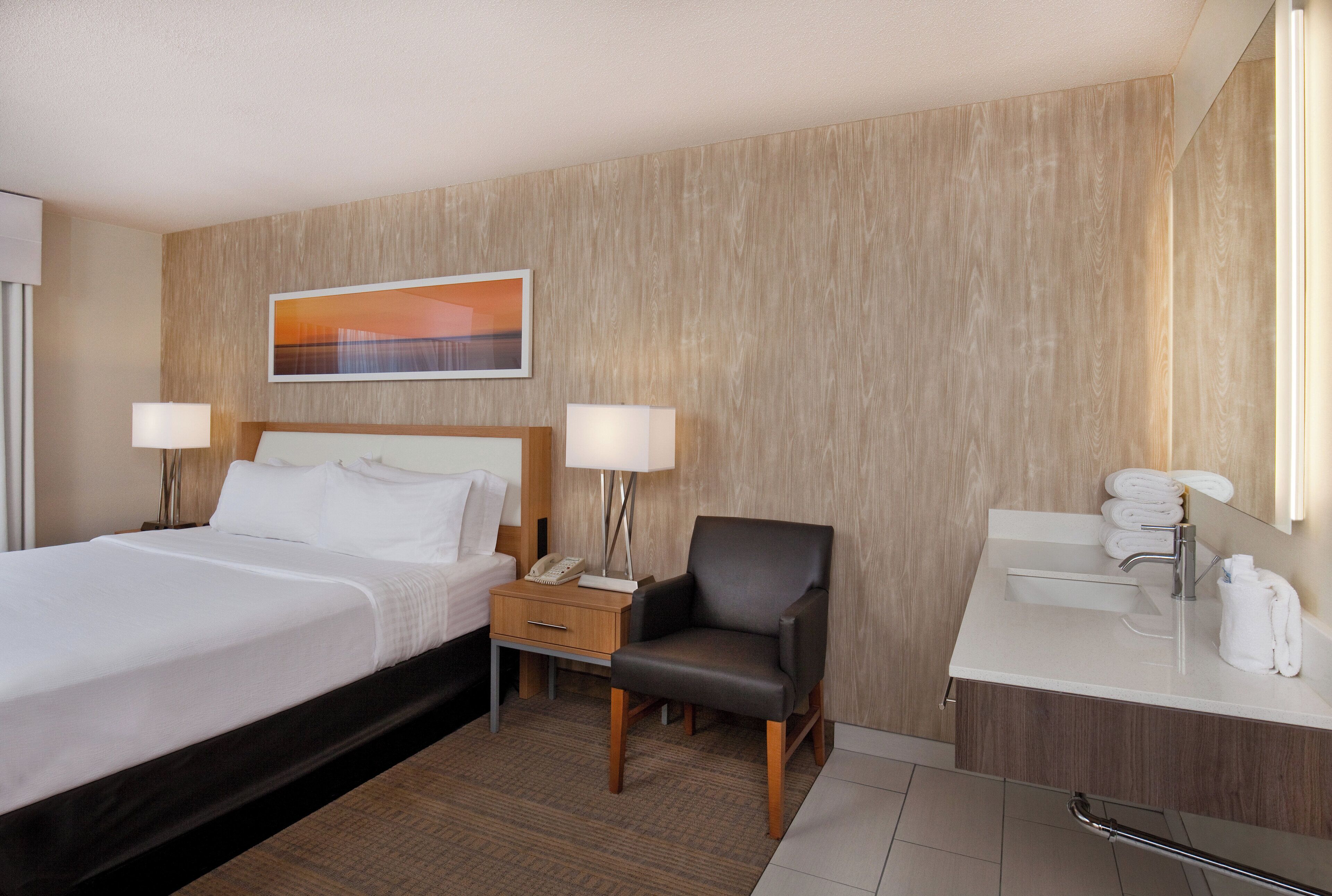 Habitación de hotel con cama, mesita de noche, silla y baño con lavamanos y espejo.