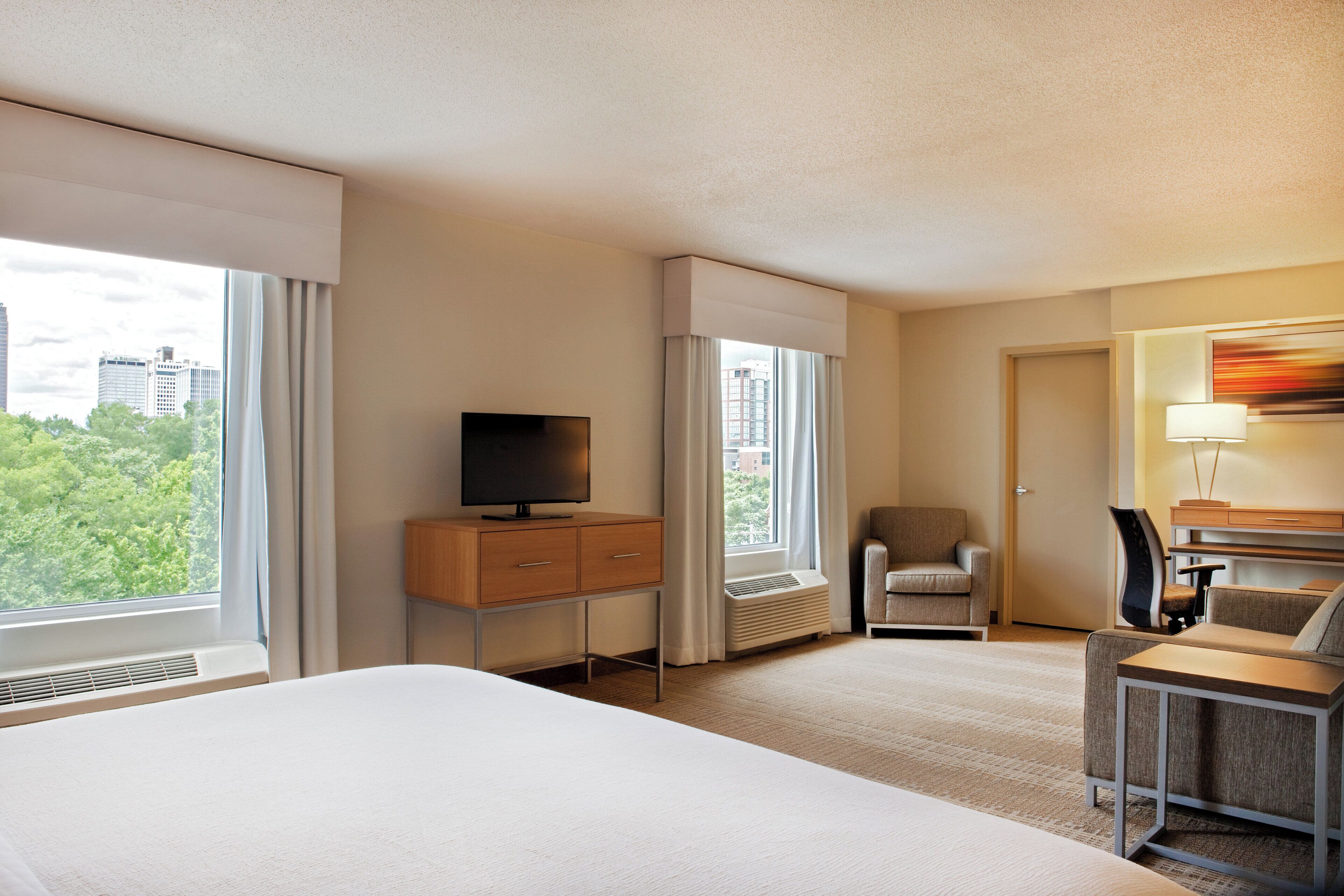 Quarto de hotel com cama, televisão, escrivaninha, cadeira e janela com vista para a cidade.