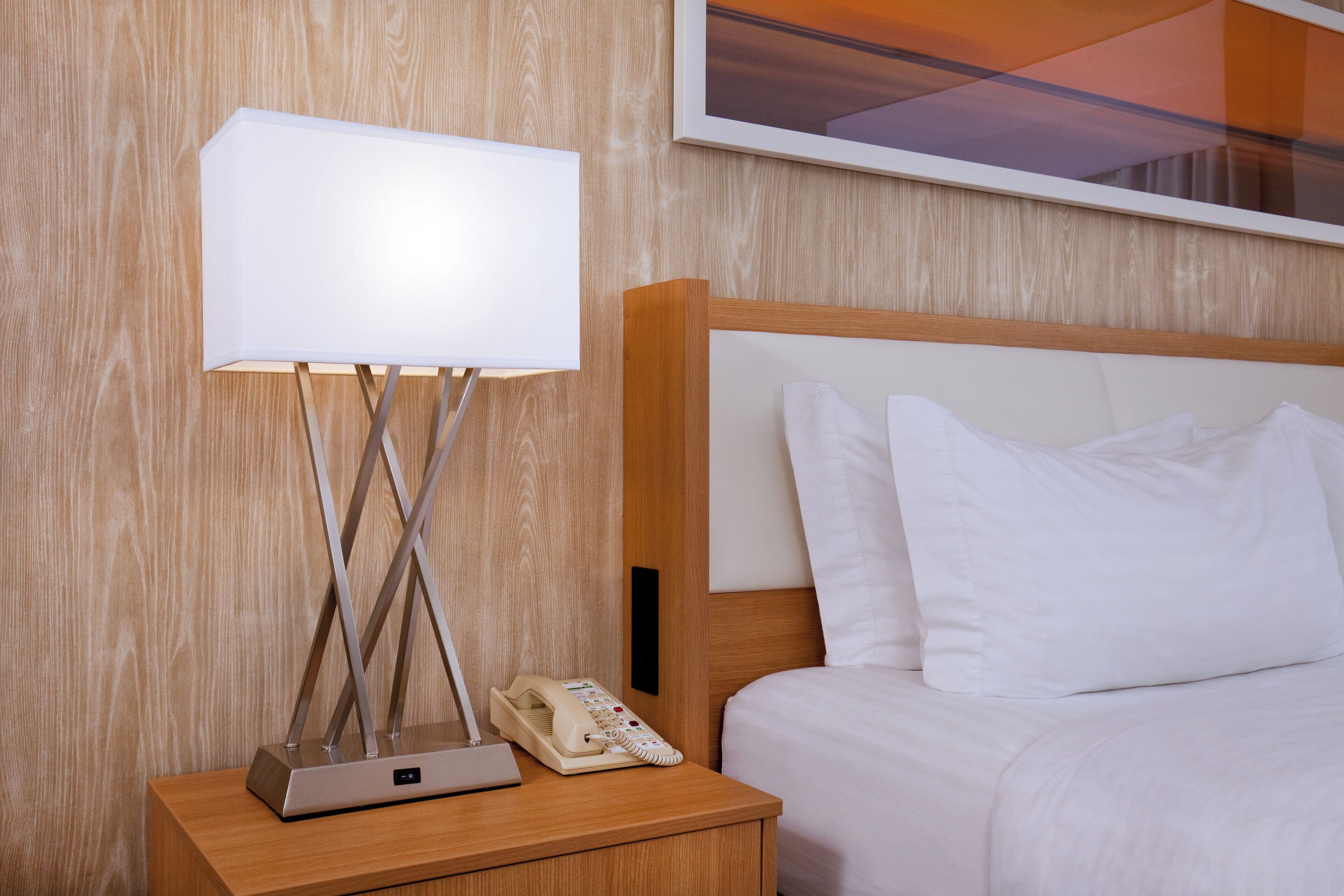 A neatly made bed with white linens, a bedside table with a lamp, a telephone, and a wall-mounted artwork.
