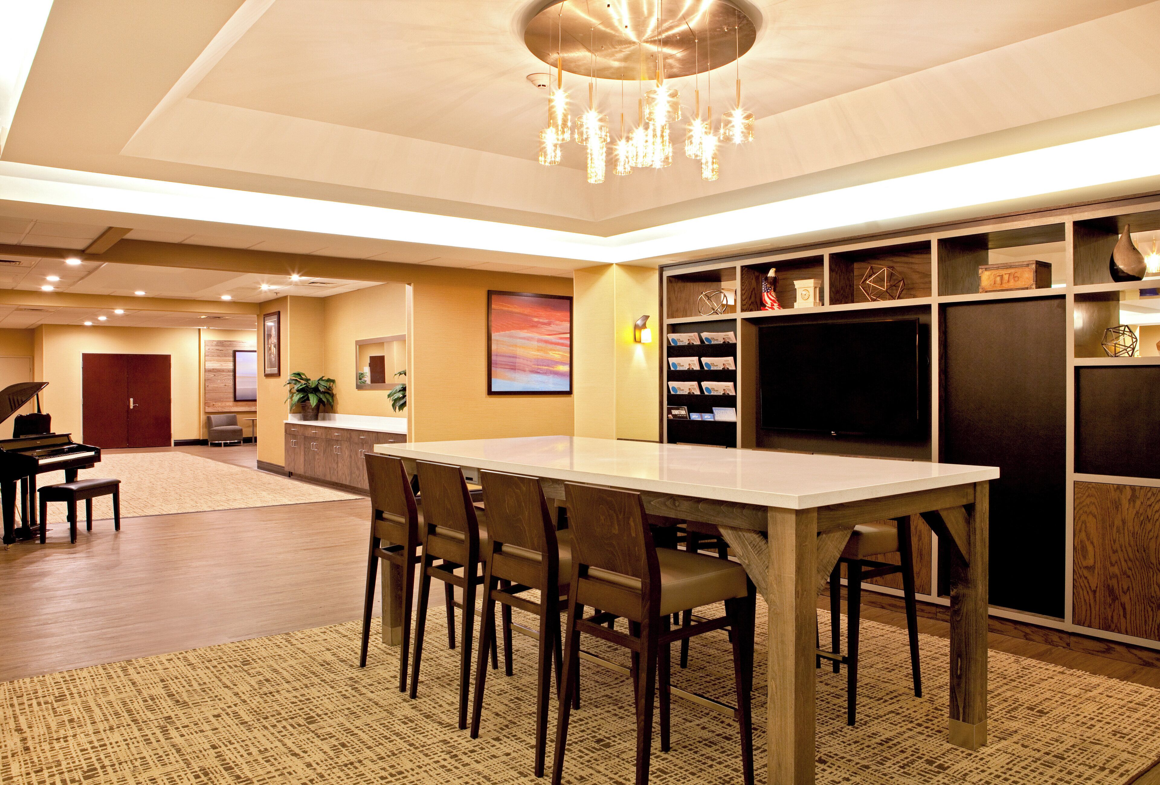 A spacious hotel lobby with a piano, a reception desk, a large dining table with chairs, and a television.