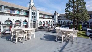 An outdoor patio area with white plastic chairs and tables, a building with arched windows and a tower, and parked cars in the background. - Castle Inn and Suites (Anaheim)