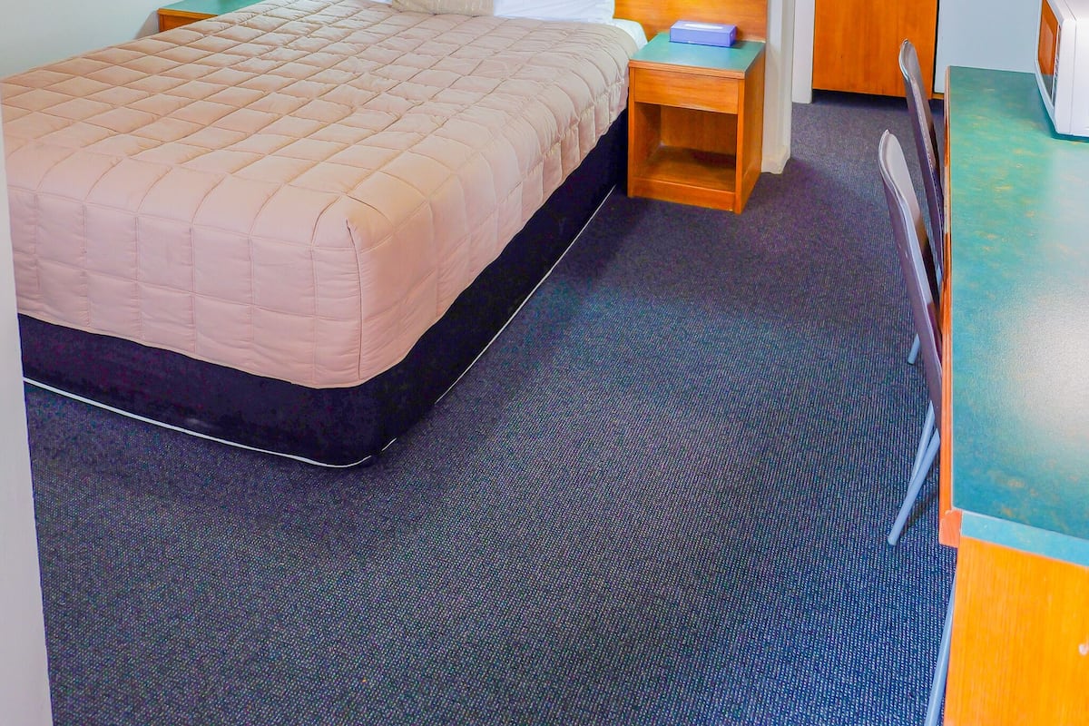 A hotel room with a bed, bedside tables, a desk, a chair, a ceiling fan, a painting, and a kitchenette.