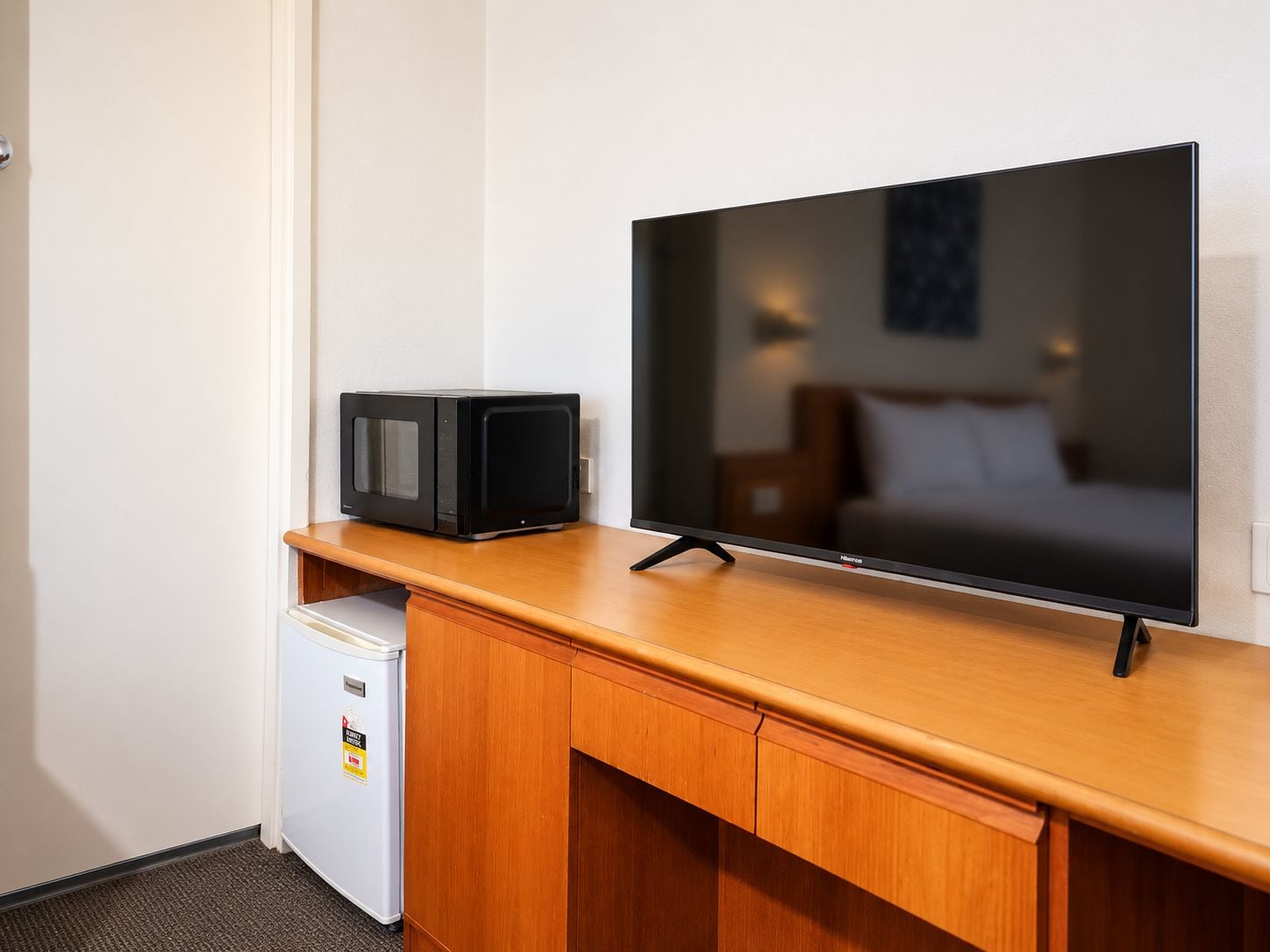 Habitación de hotel con escritorio de madera, televisor de pantalla plana, refrigerador minibar y cama con cabecera blanca.