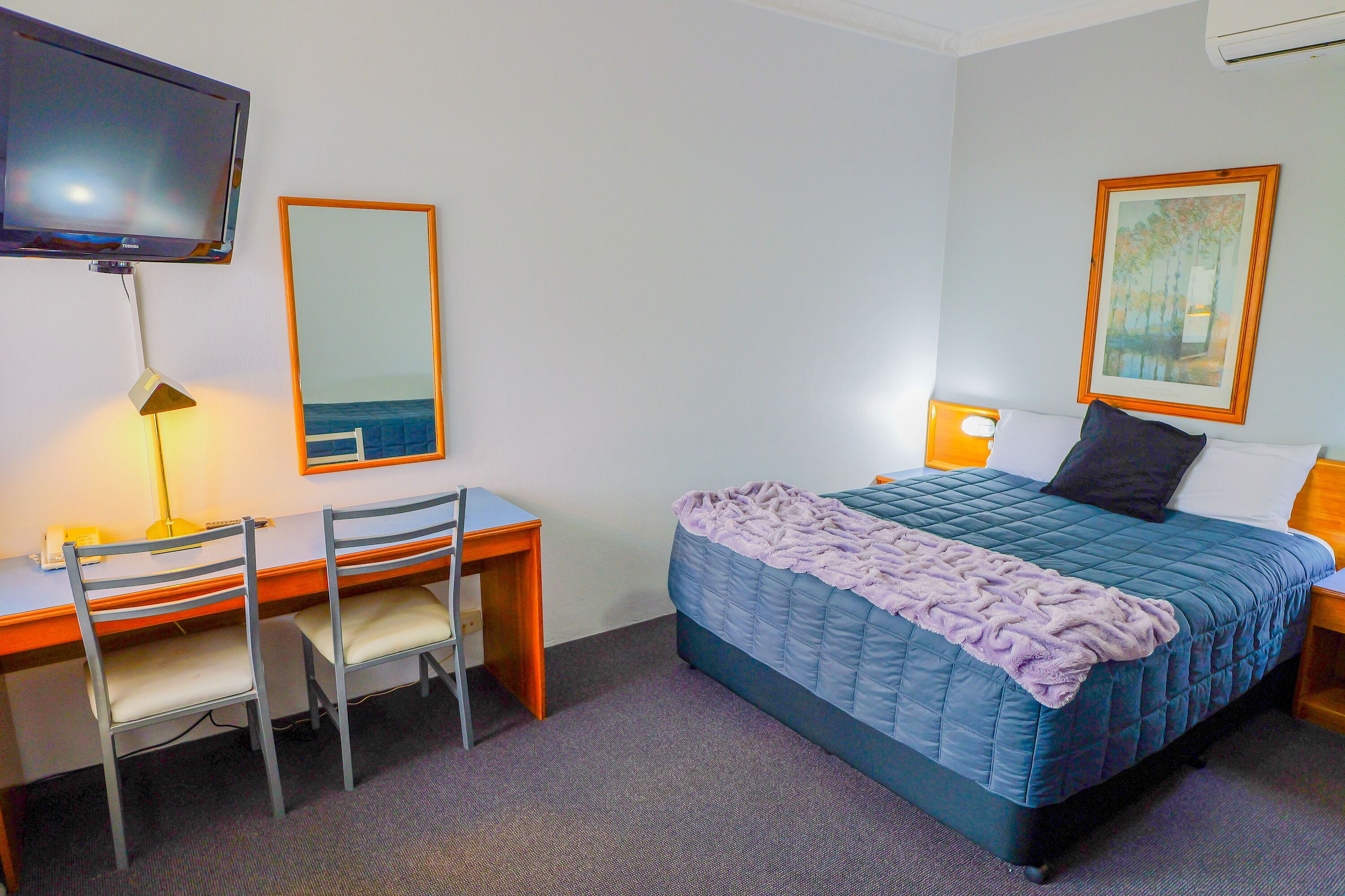 A hotel room with a bed, a desk, a chair, a TV, and a painting on the wall.