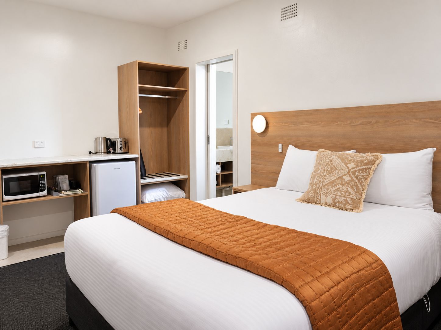 A hotel room with a bed, a desk with a microwave, a mini-fridge, and a wardrobe.