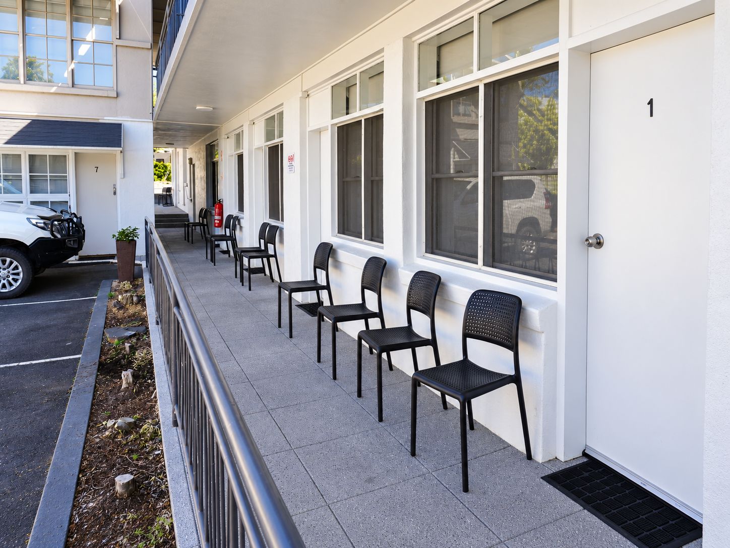 A row of black chairs outside a building with white walls and numbered doors.