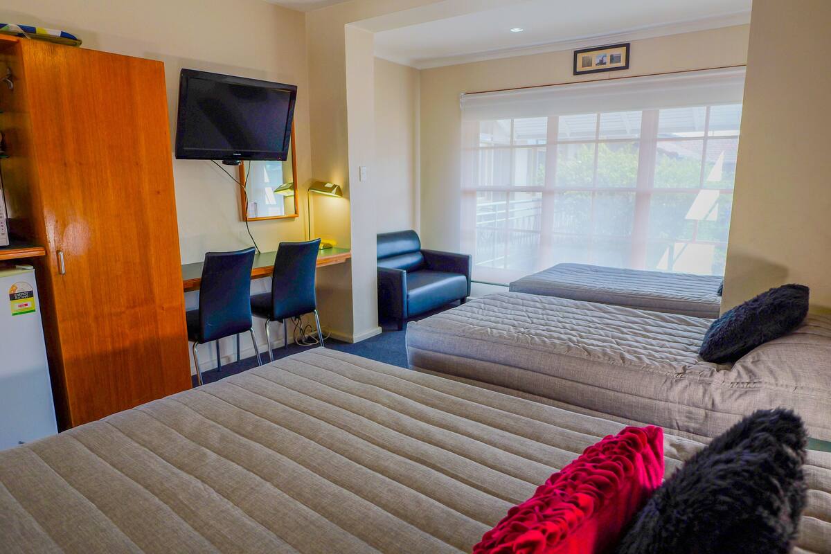 A hotel room with two beds, a desk with a computer, a TV, and a window with a view of greenery.